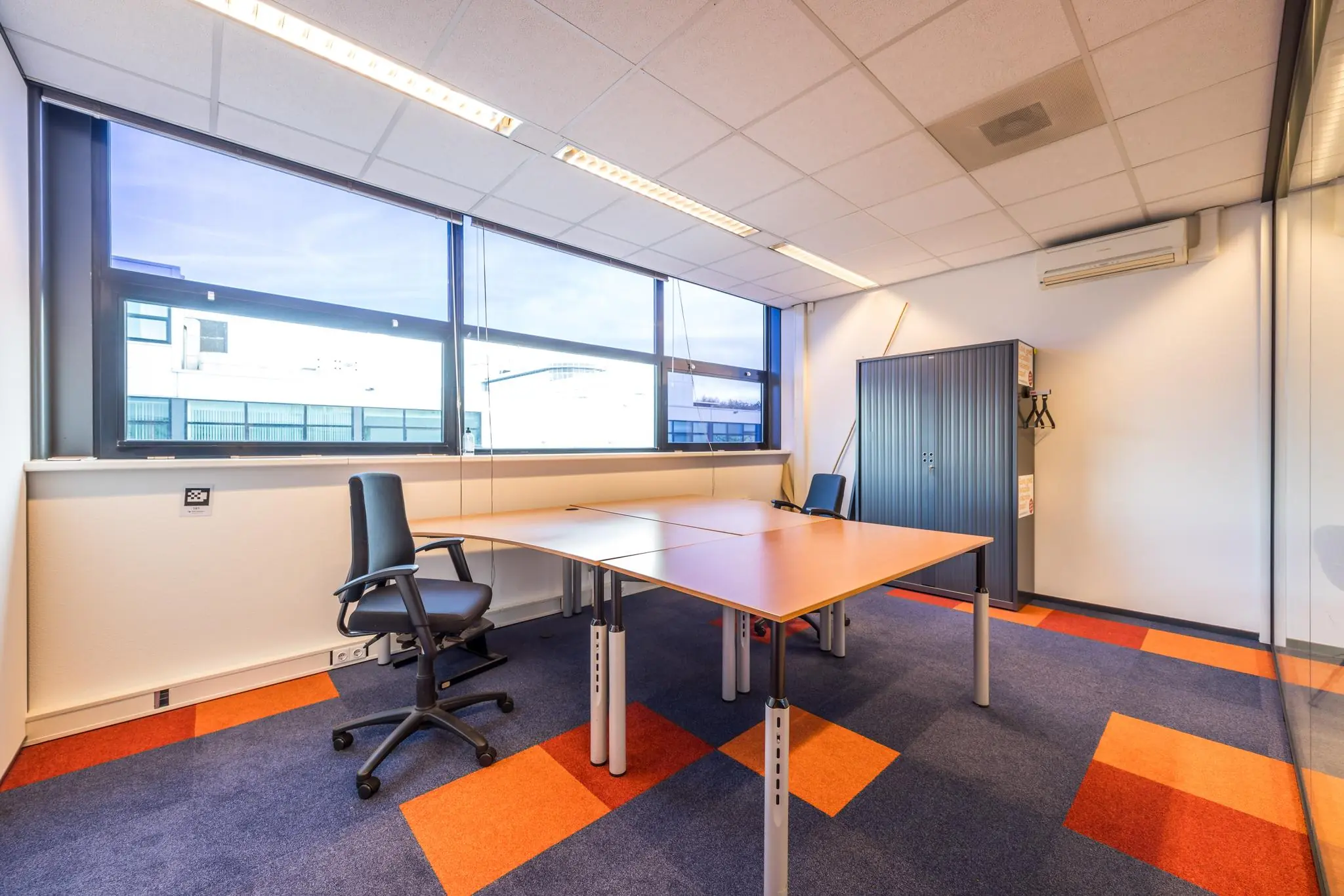 Modern kantoor met twee bureaus, ergonomische stoelen, een metalen opbergkast en grote ramen in een ruimte aan de Atoomweg.