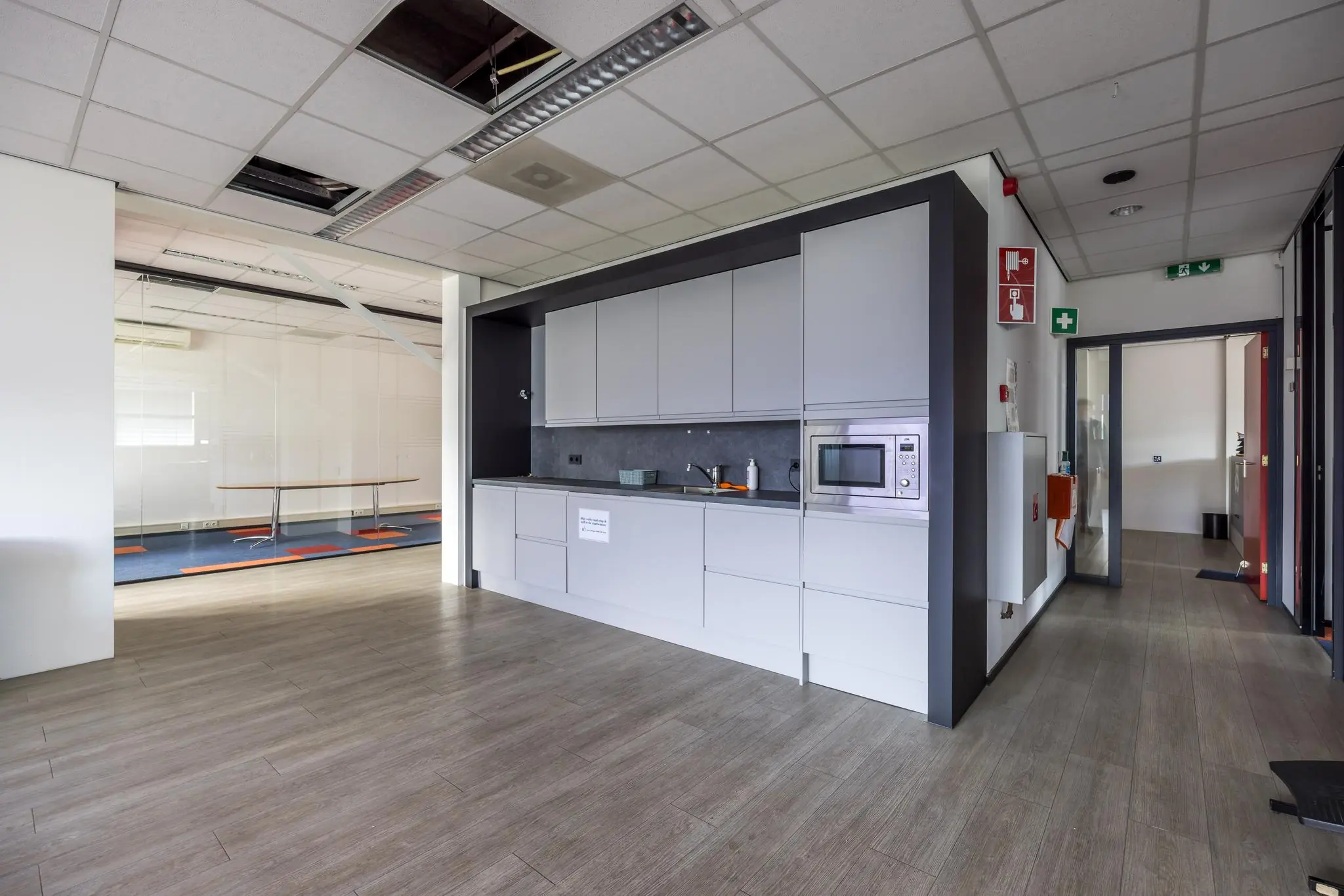 Moderne kantoorkeuken met grijze kasten, een magnetron en zicht op een vergaderruimte met glazen wanden.