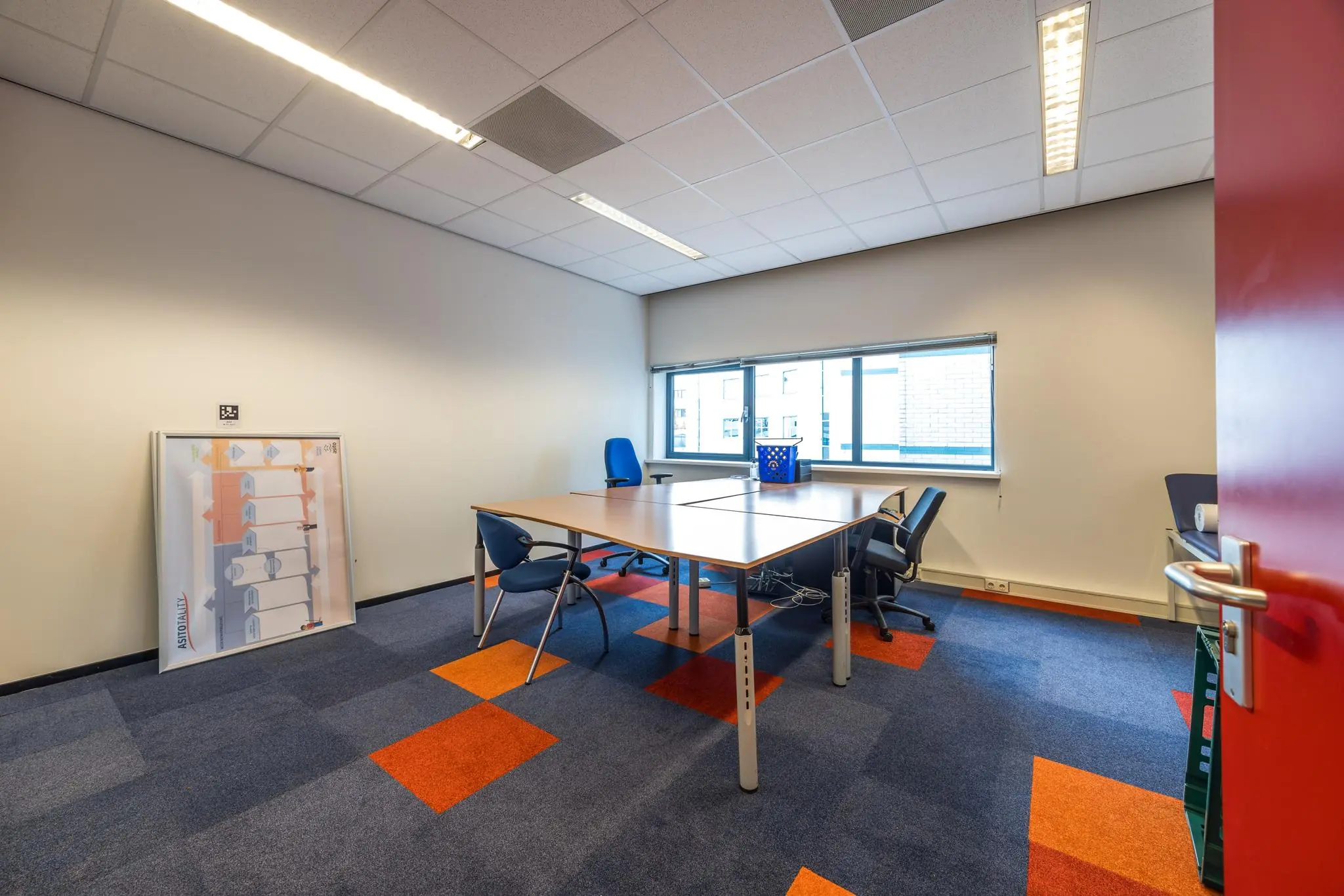 Kantoorruimte aan de Atoomweg met bureaus, stoelen, een raam en een ingelijste poster tegen de muur.