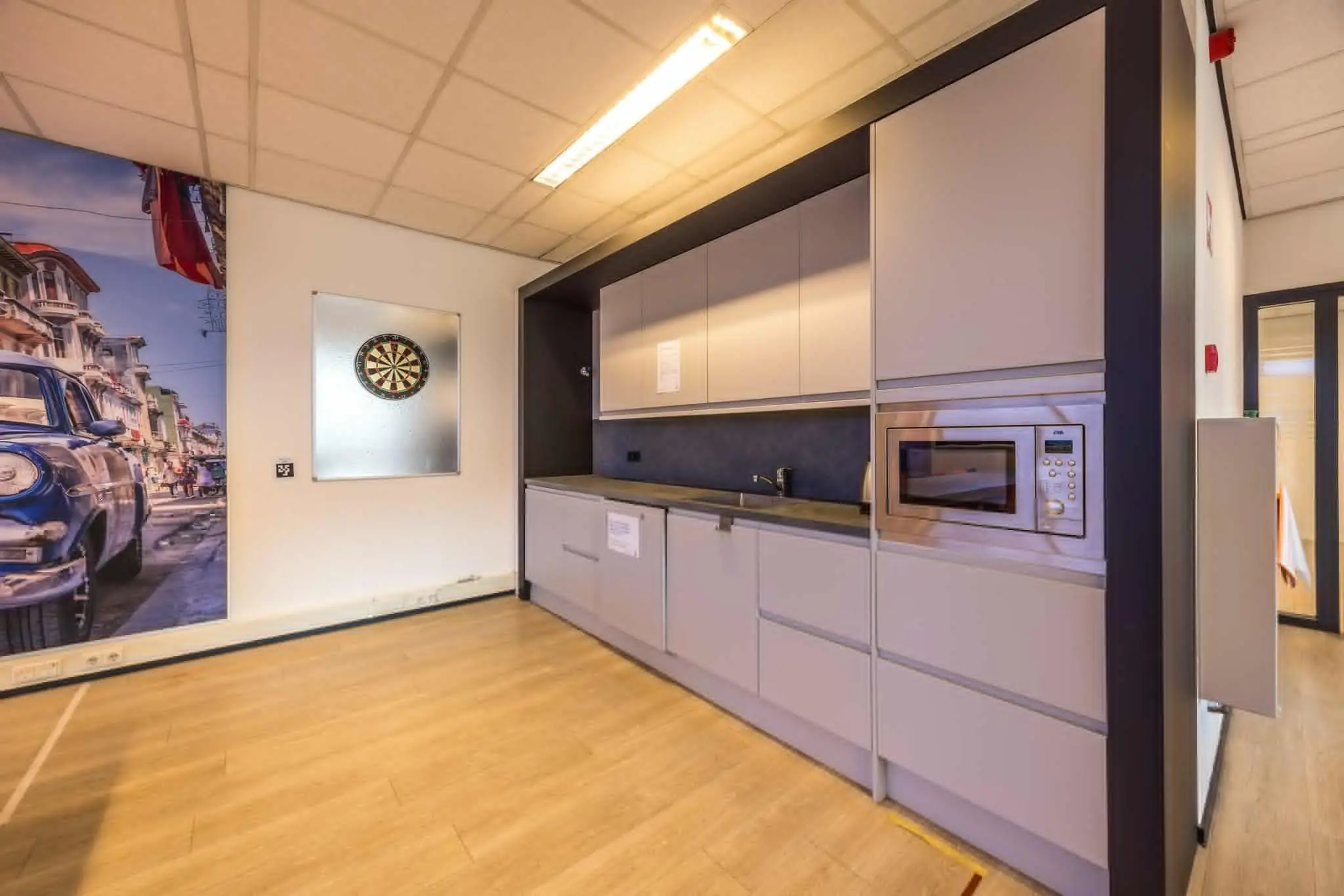 Moderne kantoorkeuken aan de Atoomweg met strakke kasten, een magnetron, dartbord aan de muur en een kleurrijke muurfoto van een straat met classic cars.