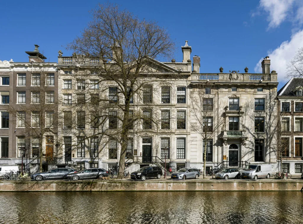 Gevelaanzicht van historische grachtenpanden aan de Herengracht in Amsterdam, met geparkeerde auto's en bomen langs het water.