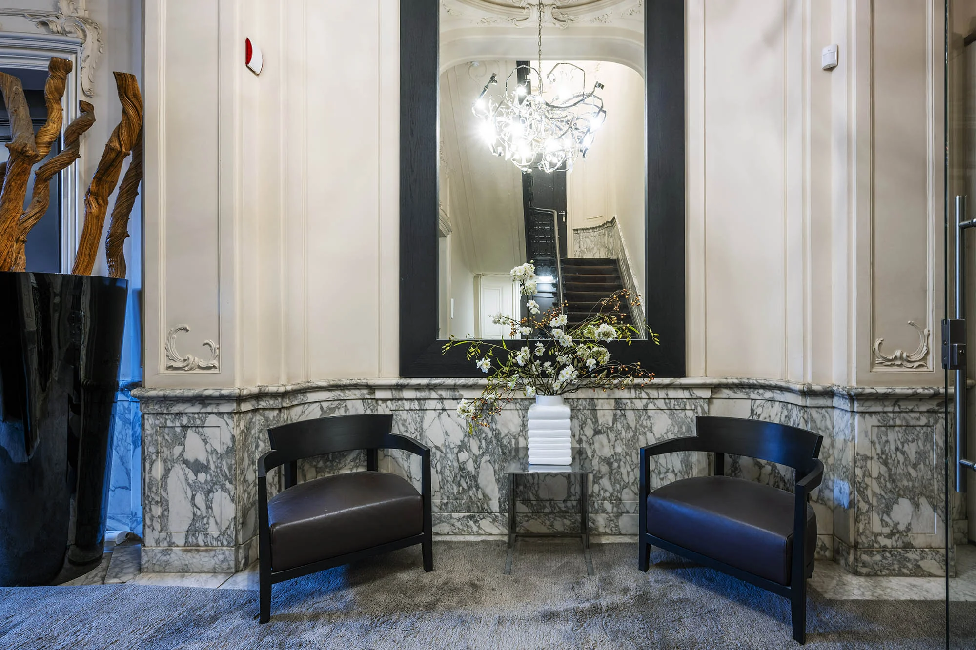 Stijlvolle entree met marmeren lambrisering, twee zwarte stoelen, een bloemenvaas en een grote spiegel die een trap reflecteert.