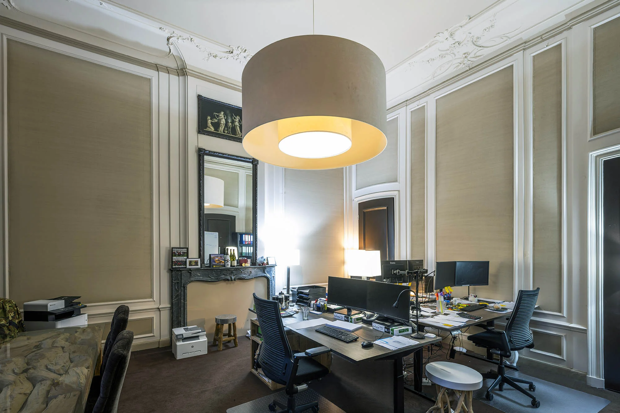 Moderne kantoorruimte aan de Herengracht met hoge plafonds, sierlijke wanddetails en een grote hanglamp boven werkplekken met meerdere beeldschermen.