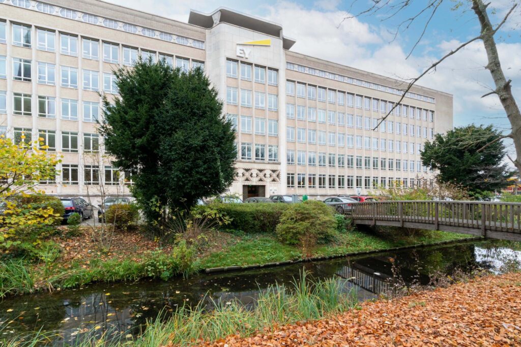 Kantoorgebouw van EY aan de Wassenaarseweg met een houten brug over een waterpartij en herfstbladeren op de voorgrond.