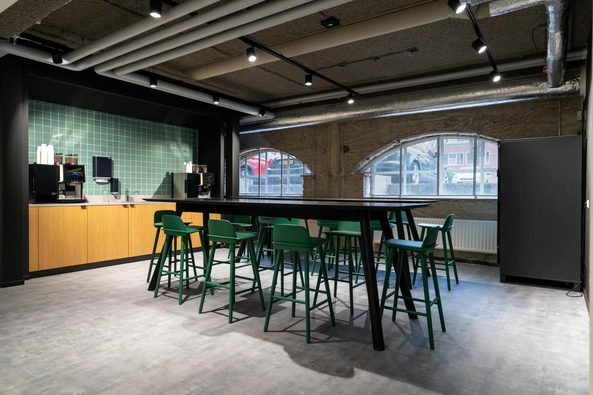 Moderne, industrieel ingerichte kantineruimte aan de Wassenaarseweg met groene barkrukken, een lange zwarte tafel en een koffiehoek met groene tegelwand.