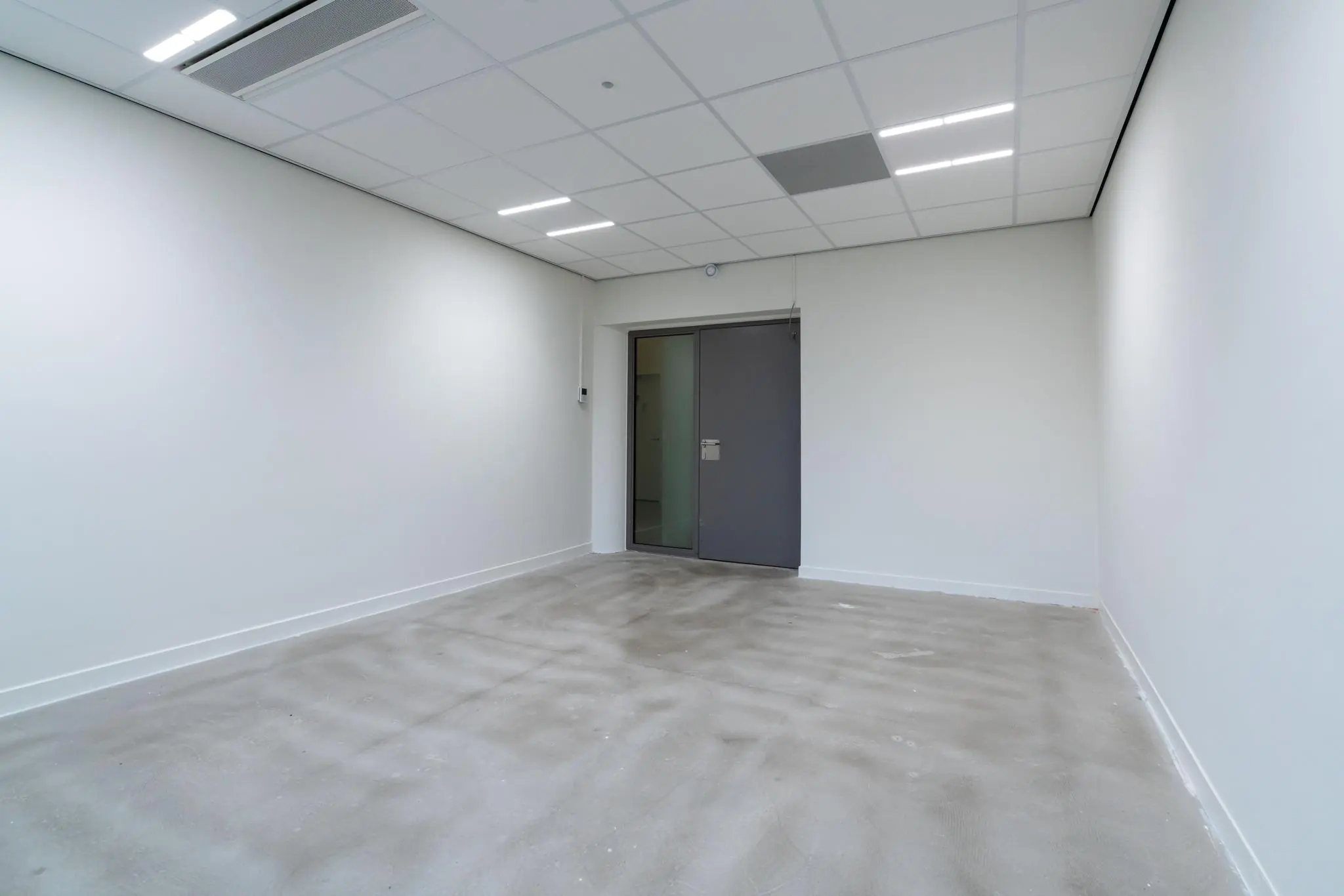 Leeg, witgeschilderd kantoor of werkkamer aan de Wassenaarseweg met systeemplafond en grijze deur.