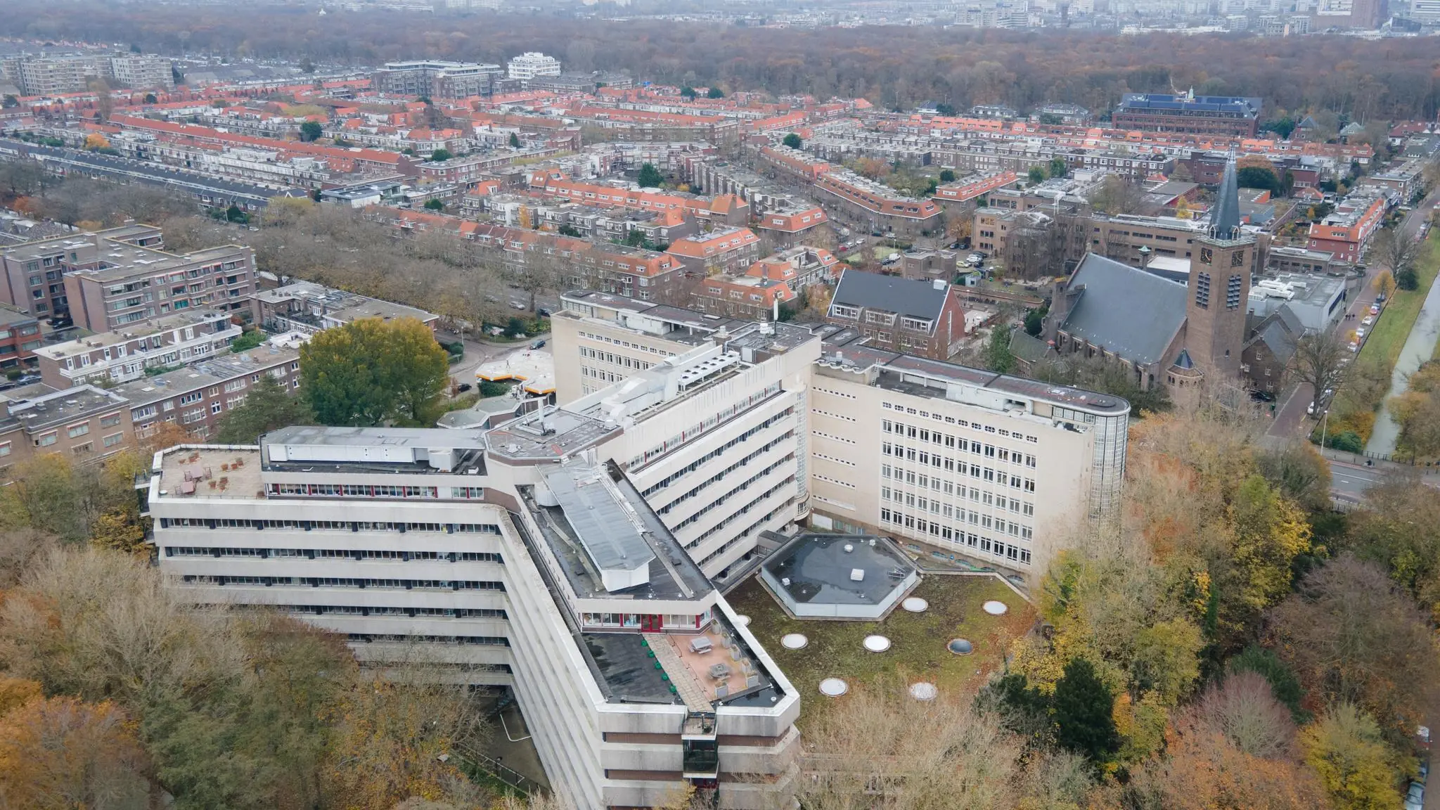 Luchtfoto van het grote gebouw aan de Wassenaarseweg in Leiden, omringd door woonhuizen, herfstbomen en een kerk.