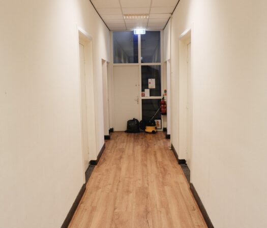 Verlichting gang met houten vloer in een pand aan de Raadhuisstraat, met schoonmaakspullen en nooduitgang aan het einde.