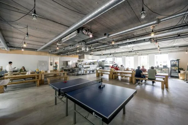 Moderne bedrijfskeuken met houten tafels, een tafeltennistafel en mensen die samen eten of werken.