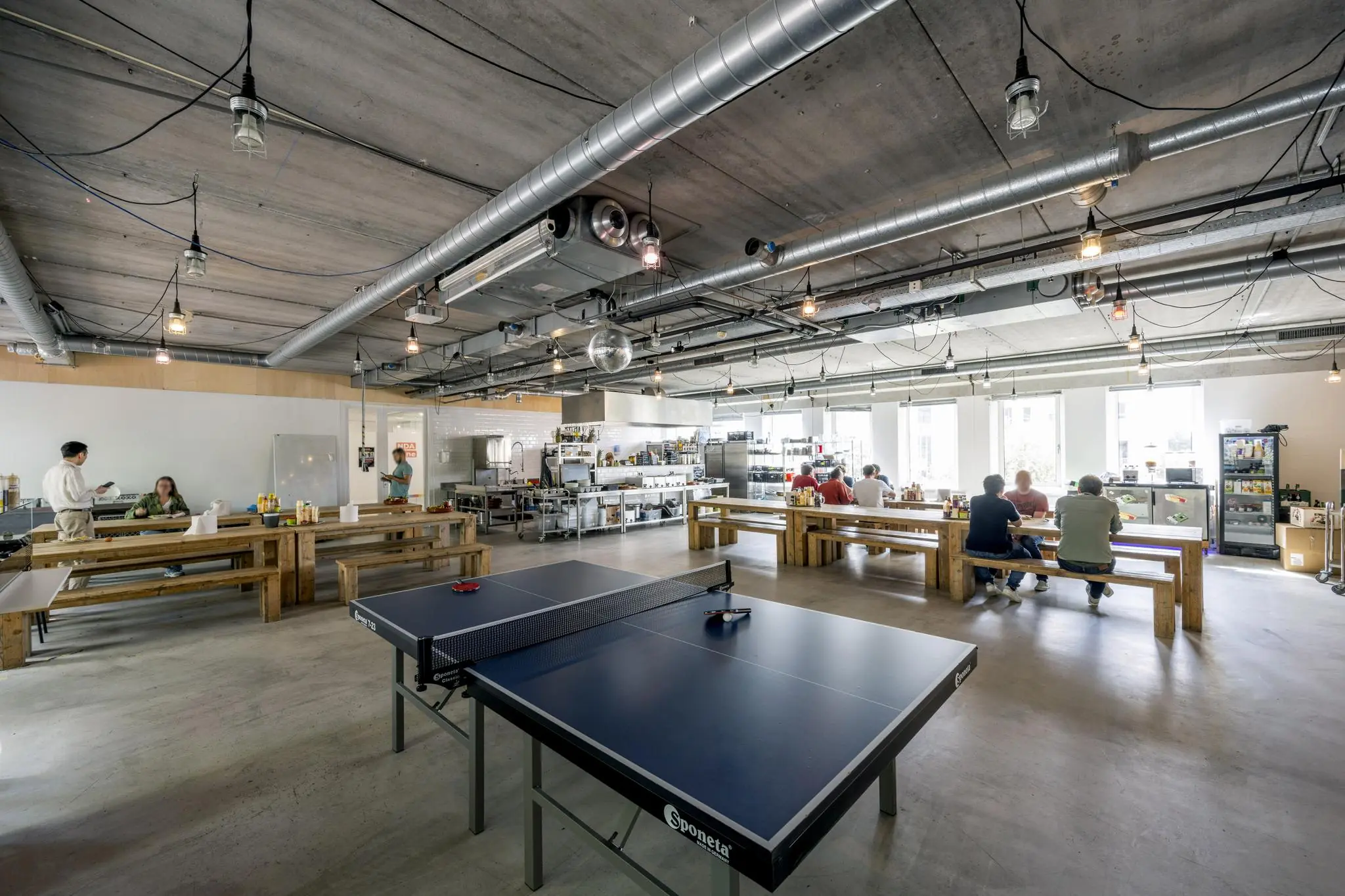 Moderne bedrijfskeuken met houten tafels, een tafeltennistafel en mensen die samen eten of werken.