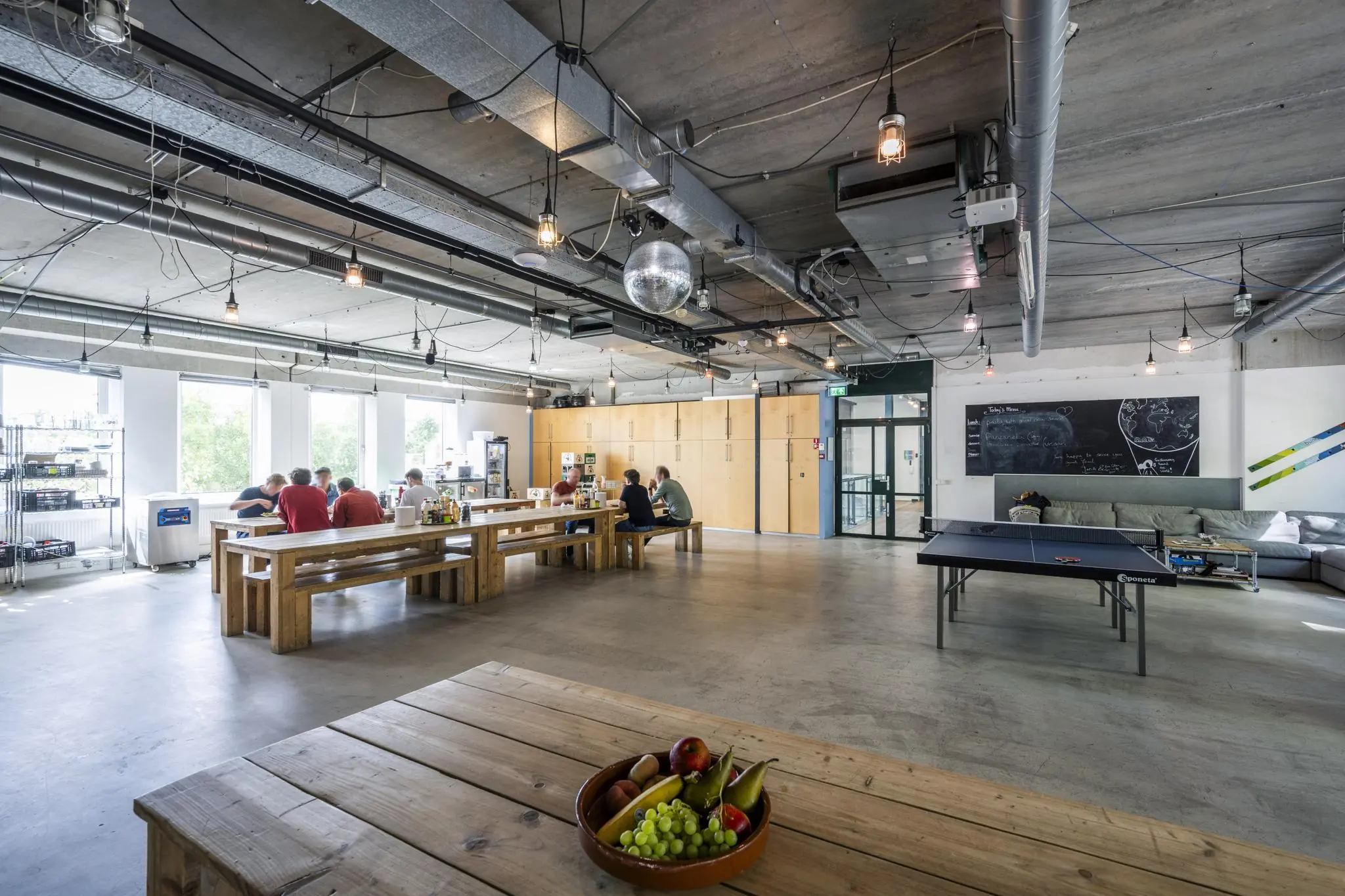 Moderne, industriële gemeenschappelijke ruimte aan de Naritaweg met houten tafels, een pingpongtafel en mensen aan het lunchen.