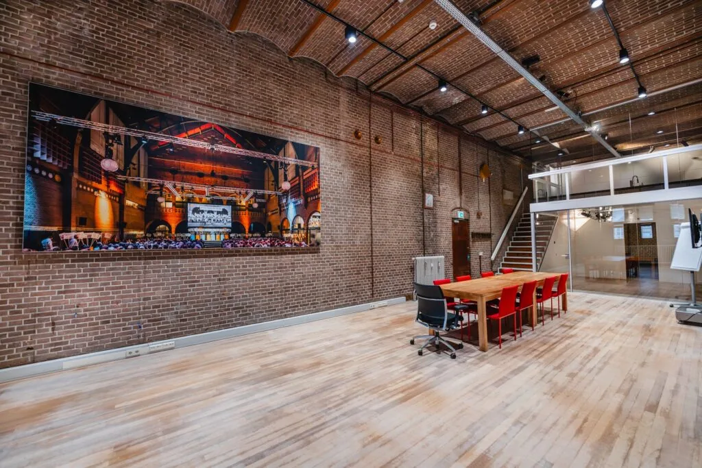 Vergaderruimte met houten vloer, bakstenen muren, moderne inrichting en een groot kunstwerk van de Beurs van Berlage aan de muur.