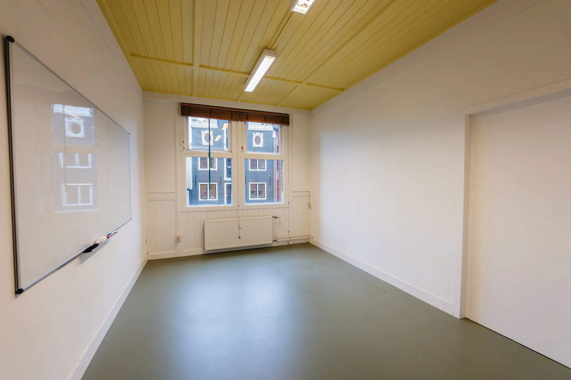 Lichte, lege kantoorruimte aan het Damrak met geel houten plafond, groot raam en whiteboard aan de muur.