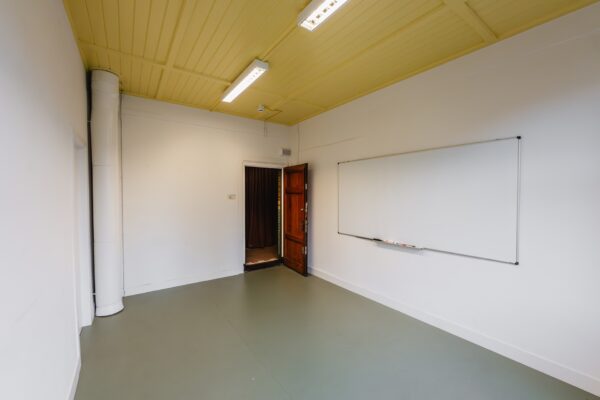 Leeg klaslokaal aan het Damrak met een geel plafond, whiteboard en open houten deur.