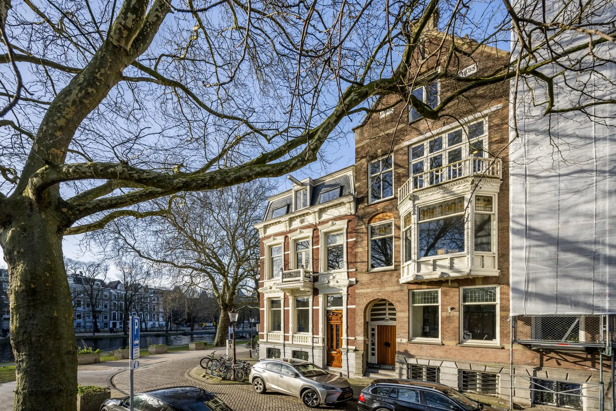Monumentale herenhuizen aan het Eerste Weteringplantsoen in Amsterdam, omgeven door bomen en geparkeerde auto's.