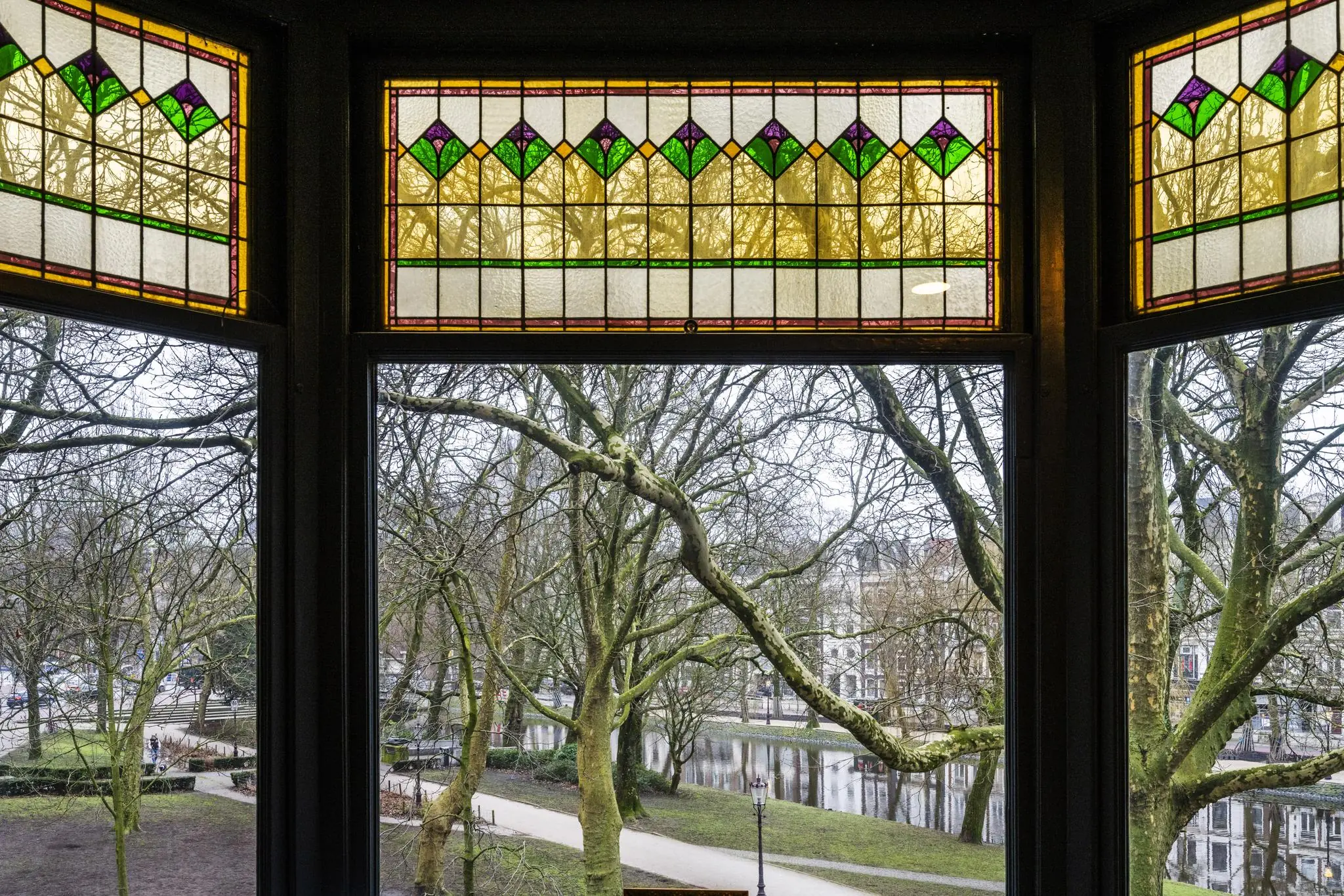Uitzicht op het Eerste Weteringplantsoen in Amsterdam door glas-in-loodramen met kleurrijk patroon.
