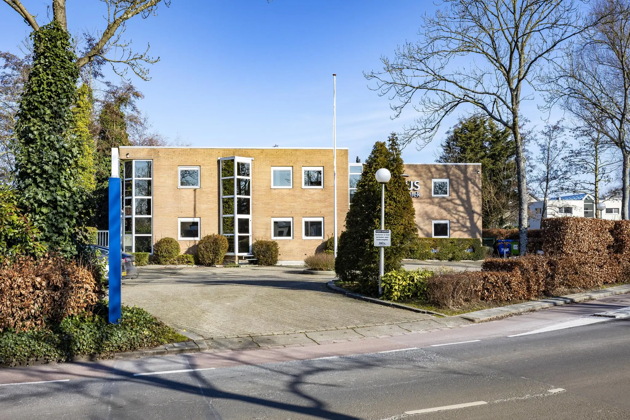 Vooraanzicht van een modern kantoorpand aan de 's-Gravenweg met een oprijlaan en omliggende bomen en struiken.