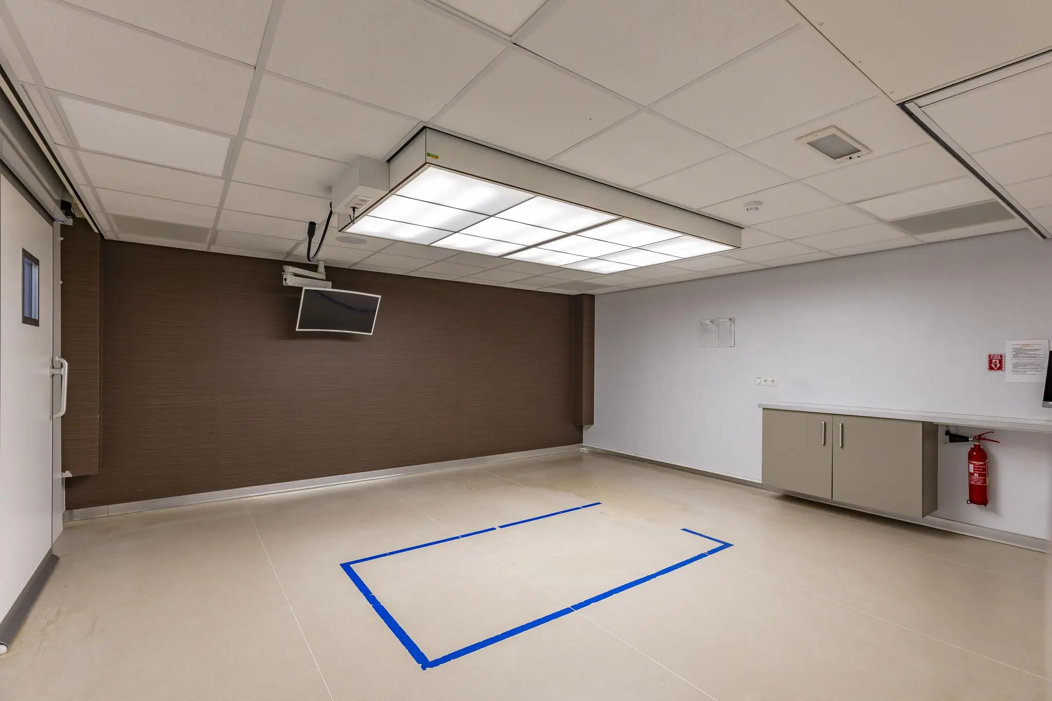 Lege kamer in een gebouw aan de ’s-Gravenweg met een gemarkeerd vlak op de vloer, plafondverlichting en een televisie aan de muur.