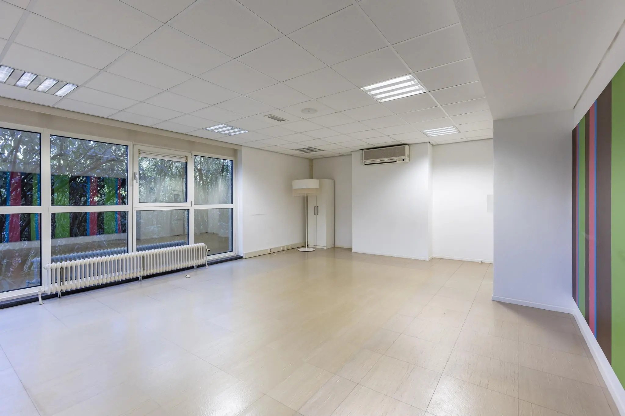 Leegstaand kantoor- of praktijkruimte aan de ’s-Gravenweg met grote ramen, een airco-unit en een gekleurde accentmuur.