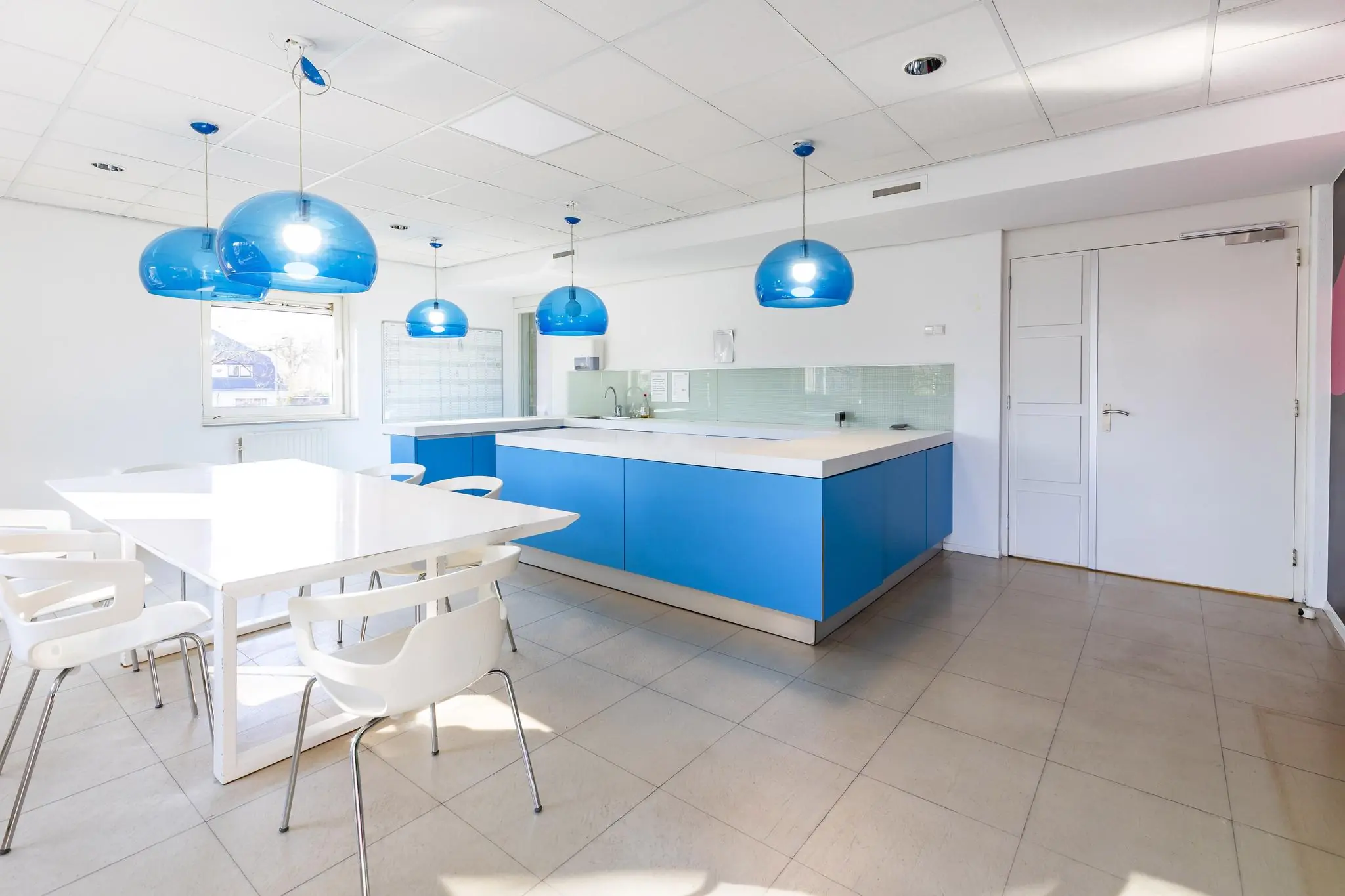 Moderne keuken met witte eettafel, blauwe keukenkasten en opvallende blauwe hanglampen.