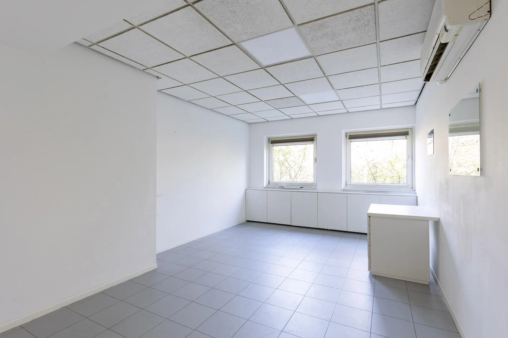 Lege, lichte kantoorruimte aan de ’s-Gravenweg met twee ramen, een systeemplafond en tegelvloer.
