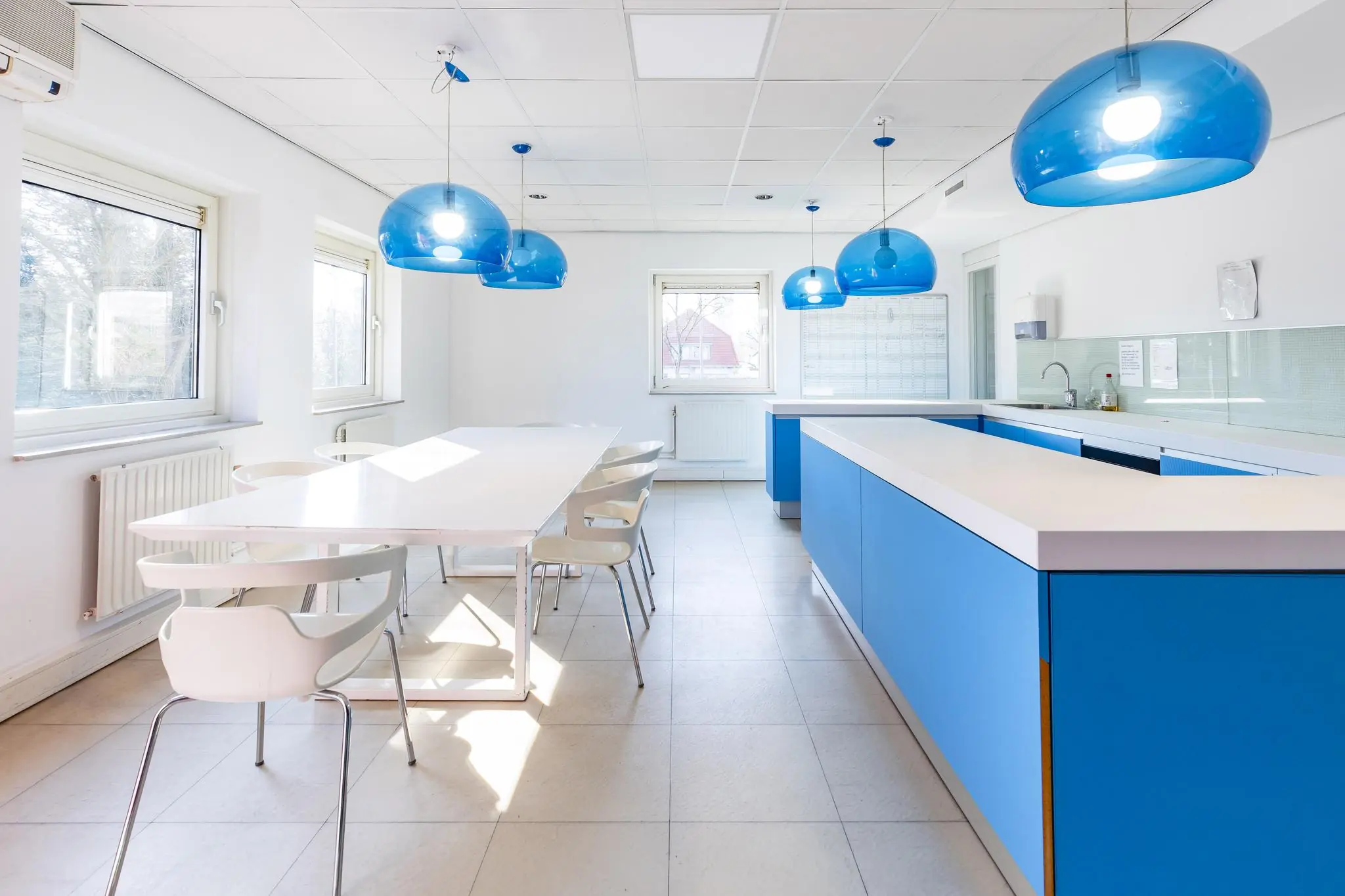 Moderne, lichte kantine met blauwe keukenkasten, witte tafels en stoelen, en opvallende blauwe hanglampen aan het plafond.