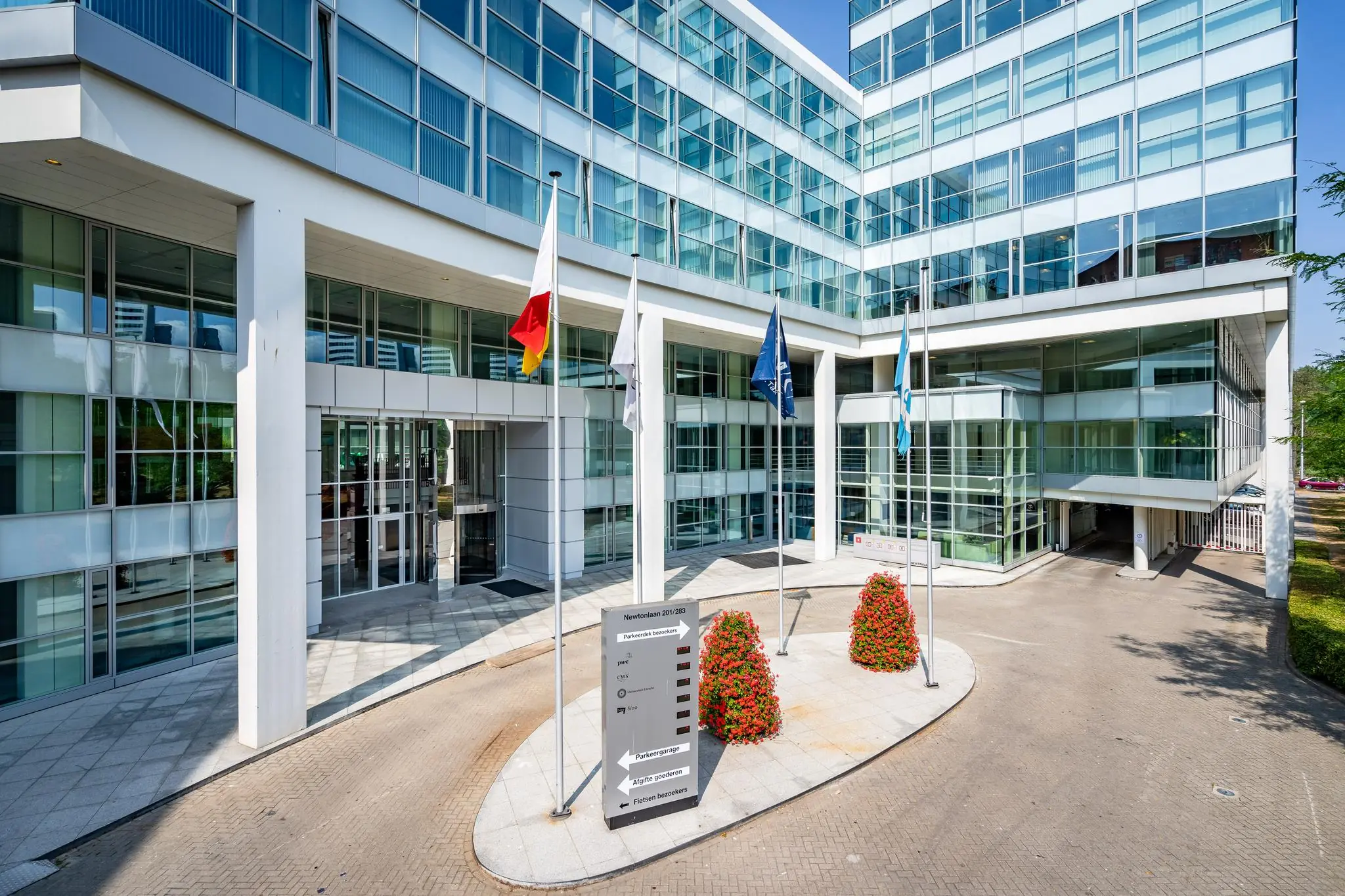 De hoofdentree van het moderne kantoorgebouw aan de Newtonlaan 201/283 met glazen gevel, vlaggen en een wegwijzer bij de oprit.