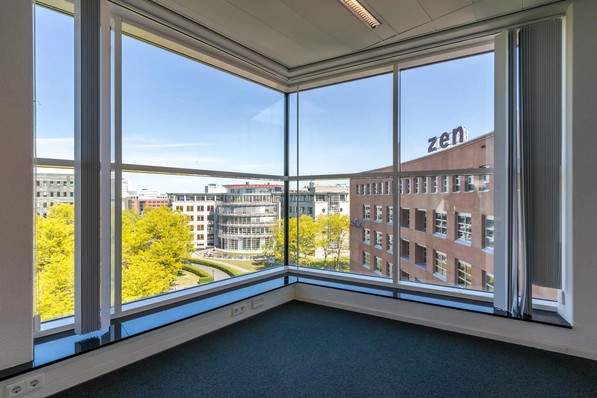 Hoek van een leeg kantoor met grote ramen en uitzicht op moderne kantoorgebouwen aan de Newtonlaan.