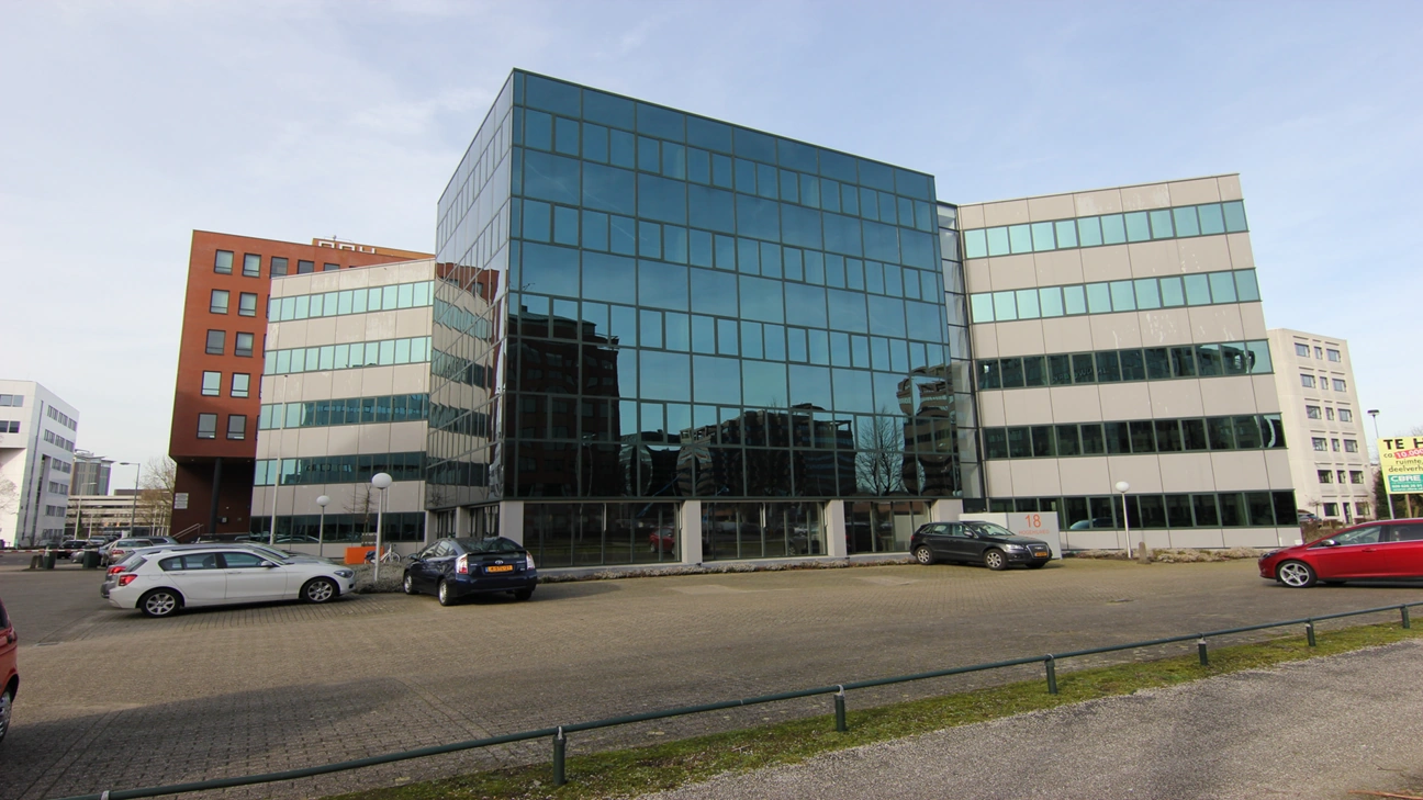 Kantoorgebouw aan de Hogehilweg 18 met spiegelende glazen gevel en parkeerterrein op de voorgrond.
