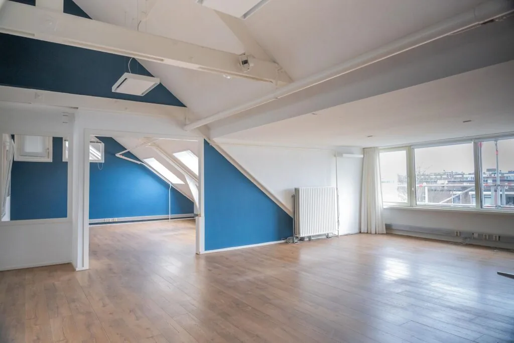 Lichte zolderruimte met houten vloer, blauwe accenten en grote ramen met uitzicht over het WG-Plein.