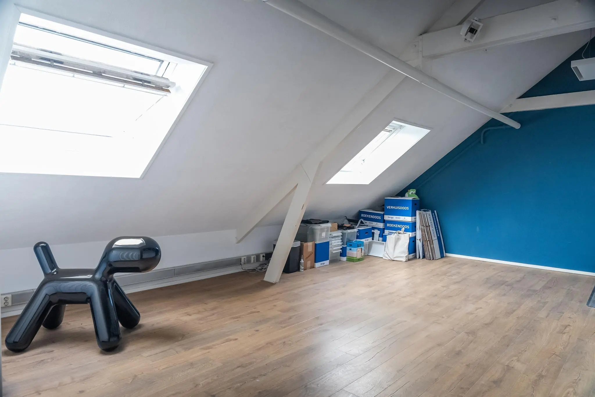 Zolderkamer met schuine daken, houten vloer, verhuisdozen en een zwart hondvormig kinderzitje bij het WG-plein.