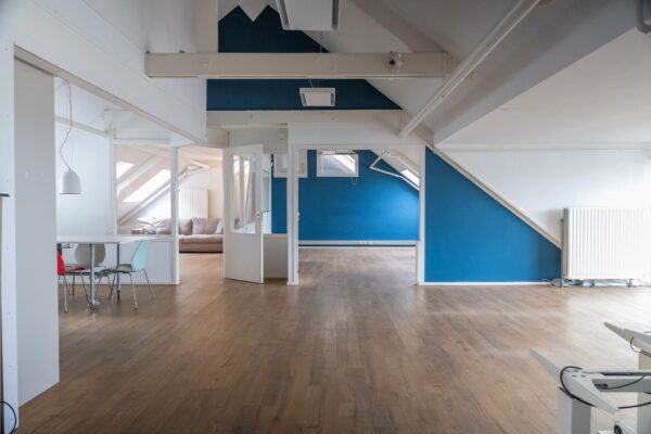 Ruime en lichte zolderverdieping met houten vloer, blauwe accentmuren en een zithoek met een beige bank.