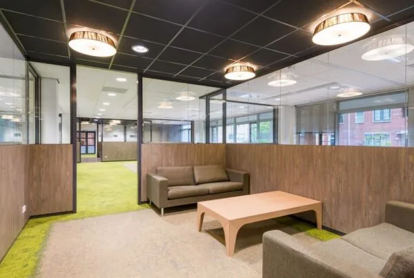 Moderne wachtruimte met glazen wanden, groene vloerbedekking en twee bruine banken rond een houten salontafel in gebouw Spui.