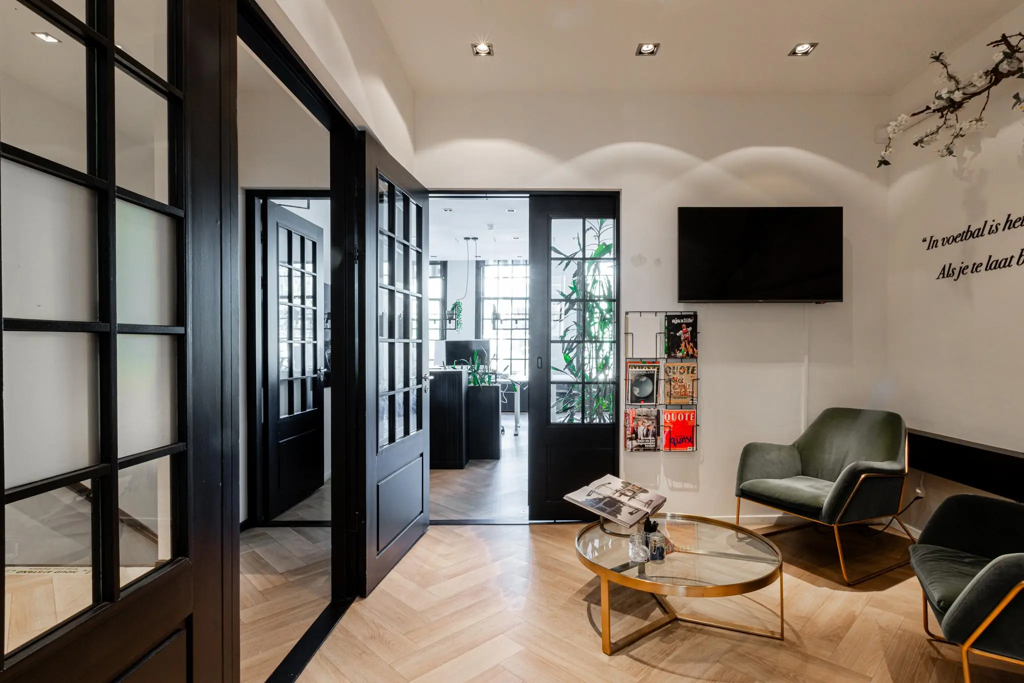 Moderne wachtruimte aan het Rokin met glazen zwarte deuren, groene stoelen, een tijdschriftenrek en een ronde glazen salontafel.