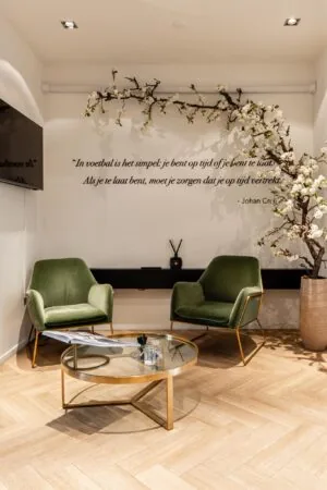 Stijlvolle wachtruimte met twee groene fauteuils, een glazen salontafel en een muurdecoratie met een citaat van Johan Cruijff.