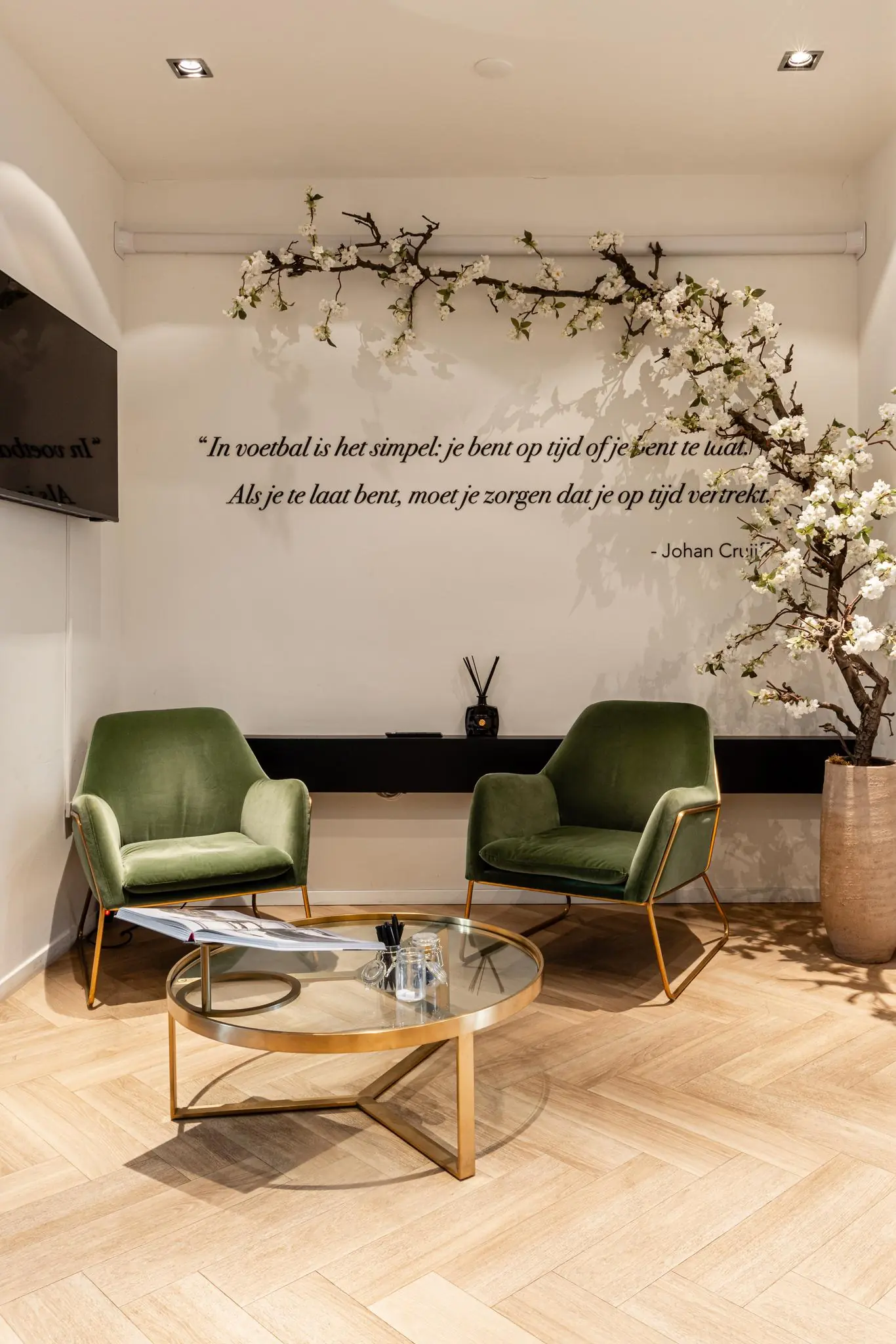 Stijlvolle wachtruimte met twee groene fauteuils, een glazen salontafel en een muurdecoratie met een citaat van Johan Cruijff.