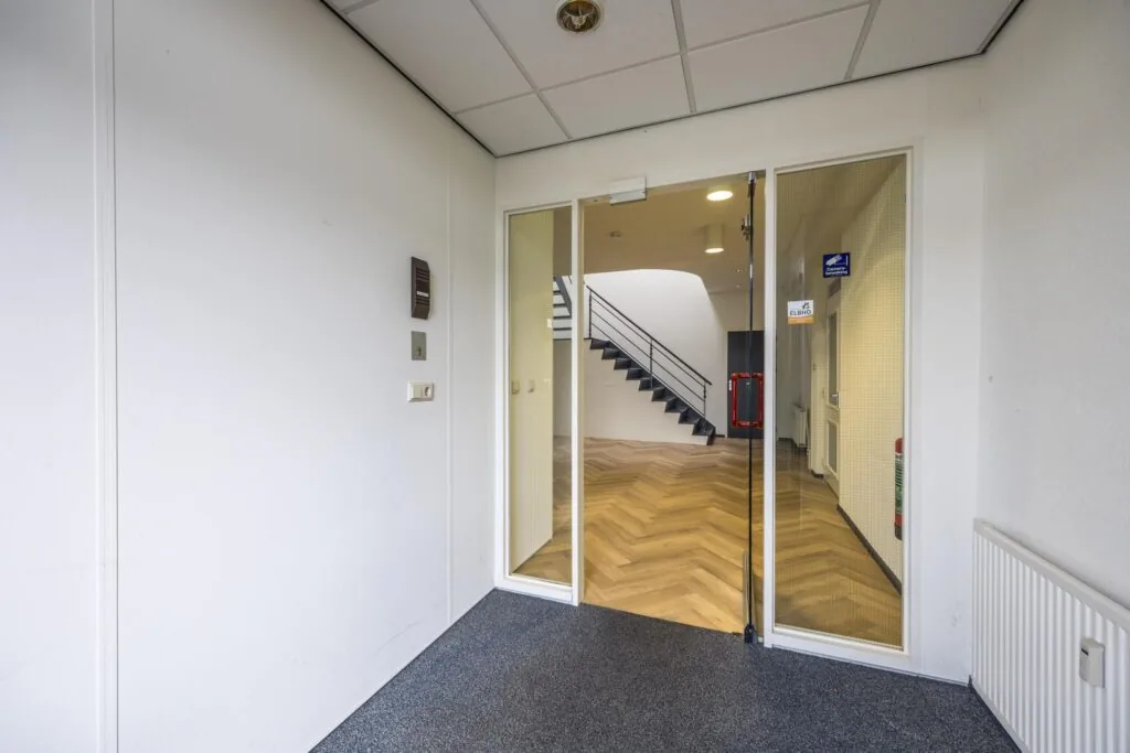 Inkomhal met glazen deur die toegang geeft tot een kantoorruimte met houten visgraatvloer en zwarte trap.