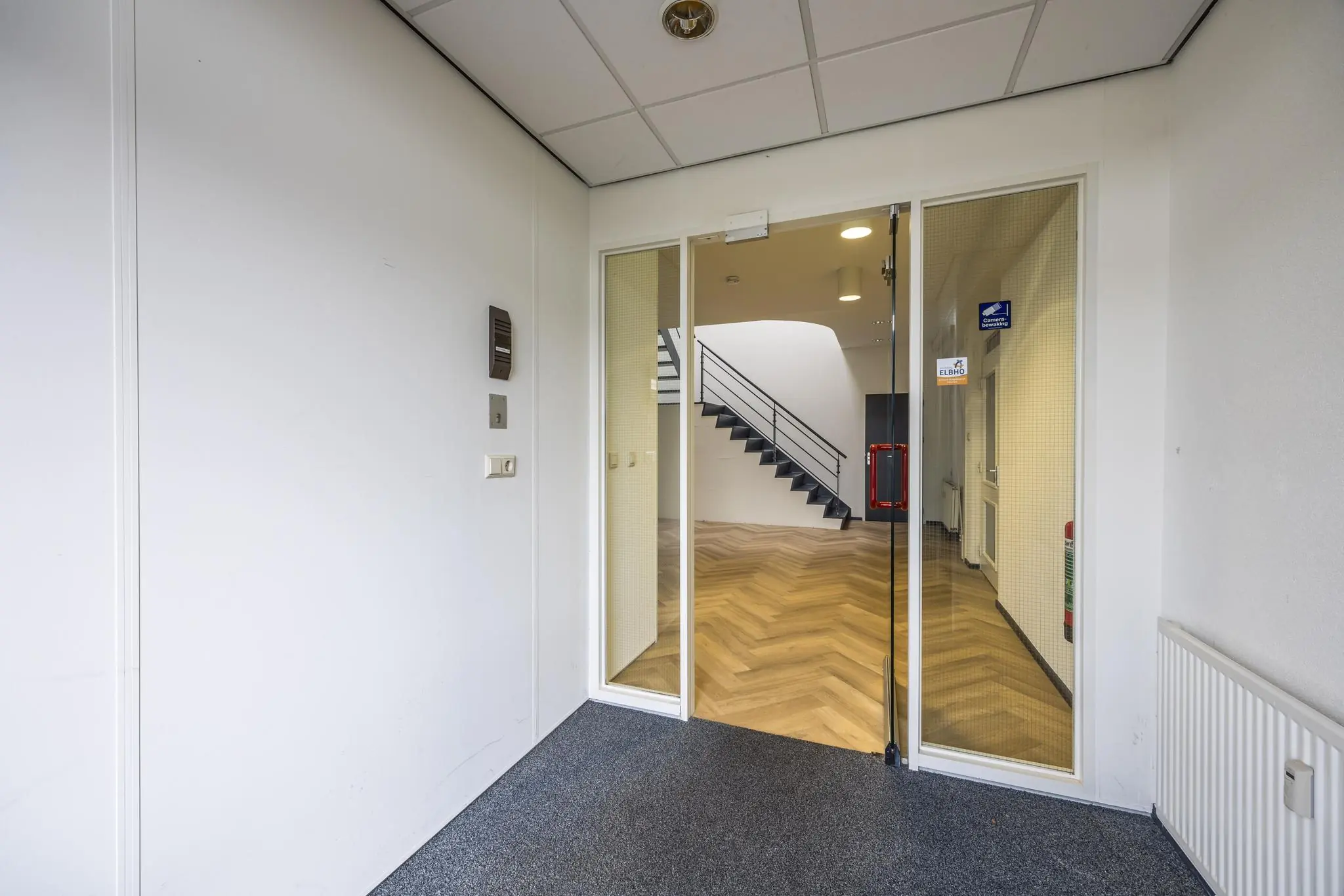 Inkomhal met glazen deur die toegang geeft tot een kantoorruimte met houten visgraatvloer en zwarte trap.