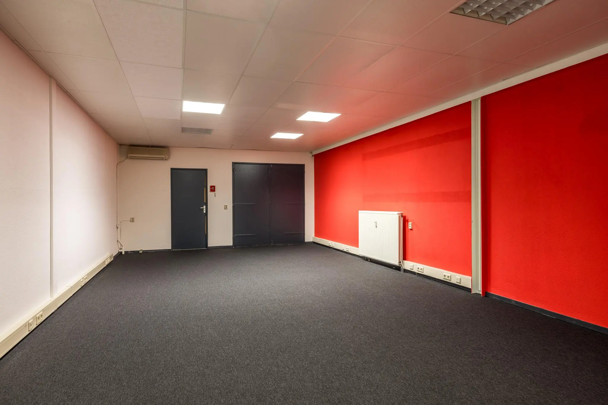Lege kantoorruimte met grijze vloerbedekking, rode accentmuur en dubbele grijze deuren aan de achterzijde.
