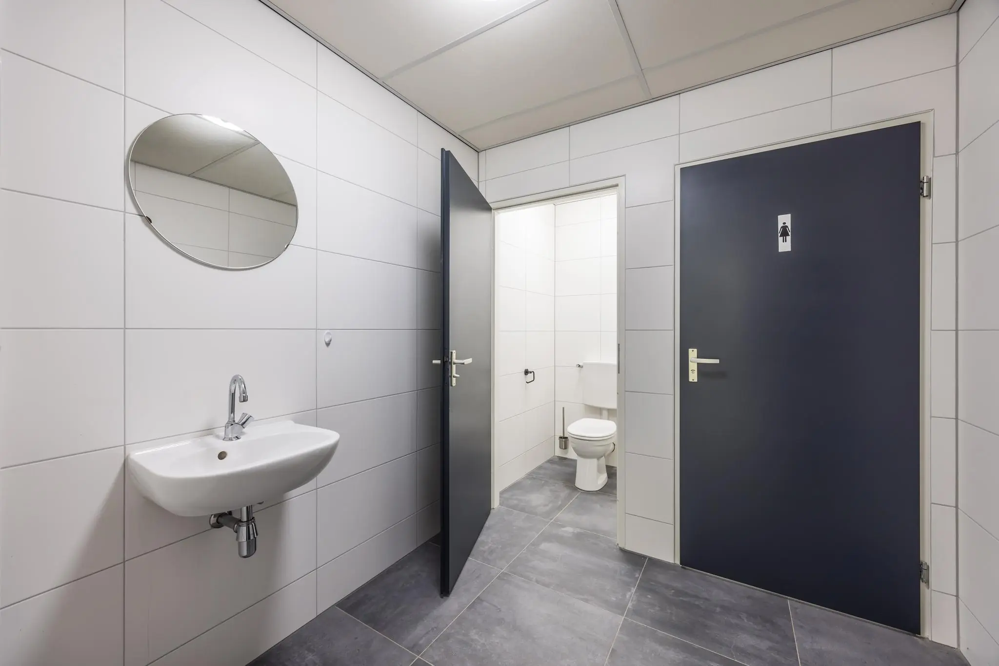 Modern en strak ingericht openbaar toilet met wastafel, spiegel en twee toiletruimtes, waaronder één voor dames.