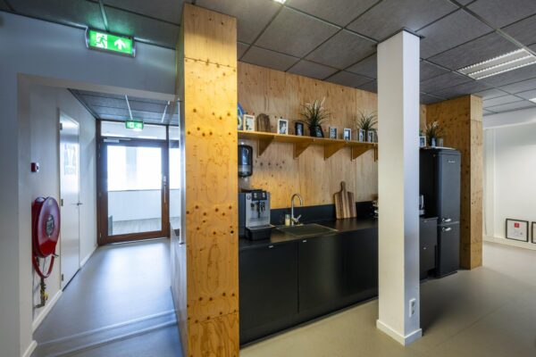 Moderne pantry in een kantoor aan de De Ruijterkade met houten wand, zwarte keukenkasten en een koffiezetapparaat.