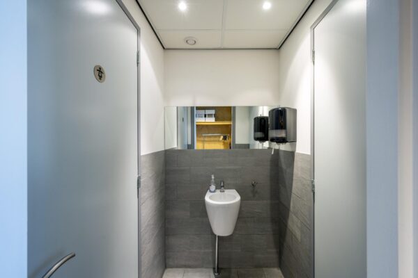Moderne, betegelde toiletruimte met een urinoir, spiegel en twee zeepdispensers aan de De Ruijterkade.