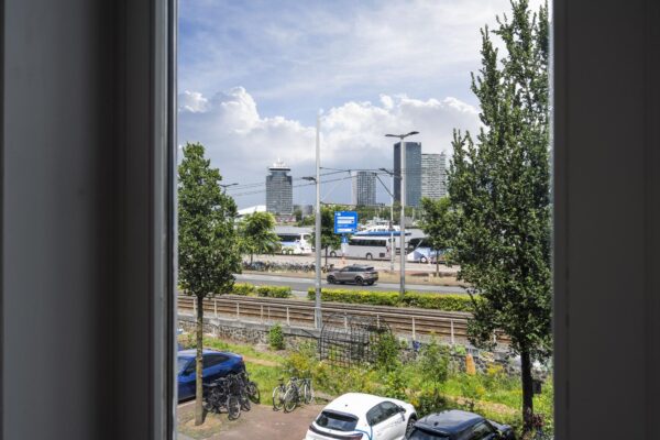 Uitzicht vanuit een raam op De Ruijterkade in Amsterdam met trambaan, auto's, fietsen en moderne hoogbouw op de achtergrond.