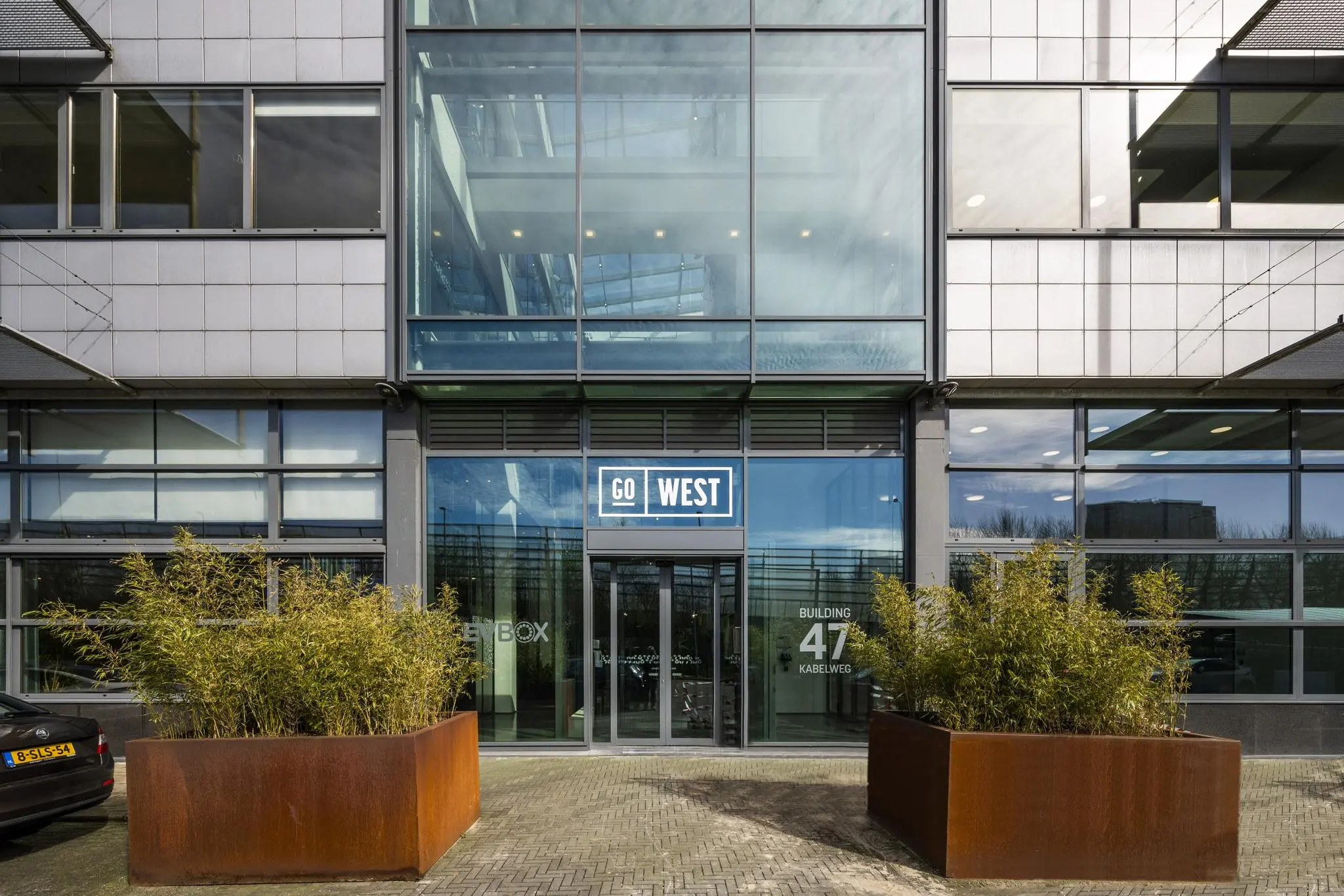 Ingang van het kantoorgebouw "Go West" aan de Kabelweg 47 met grote glazen gevel en plantenbakken.