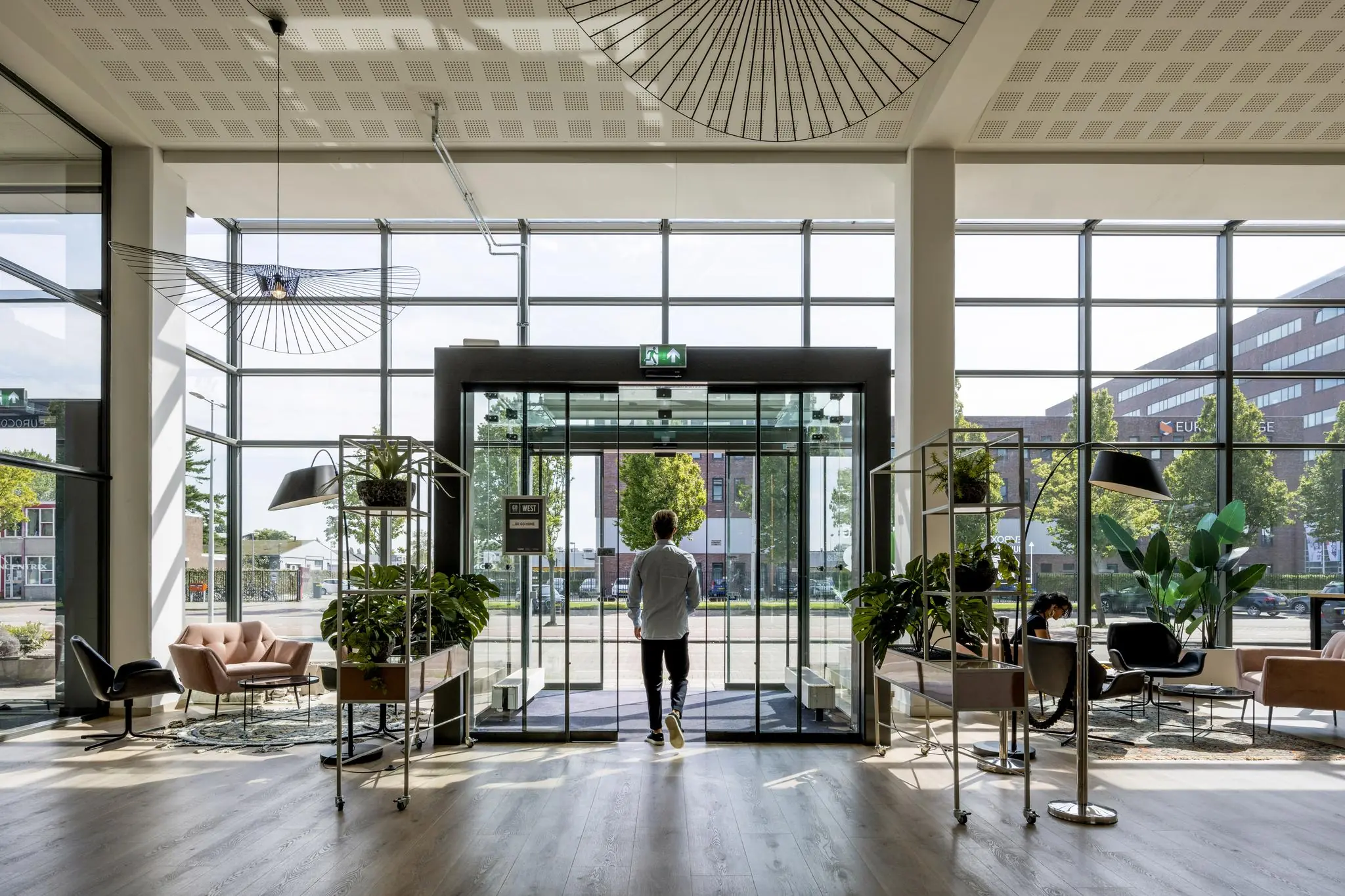 Een man loopt een modern en licht kantoor- of bedrijfsgebouw uit aan de Kabelweg, omgeven door planten en designmeubels.