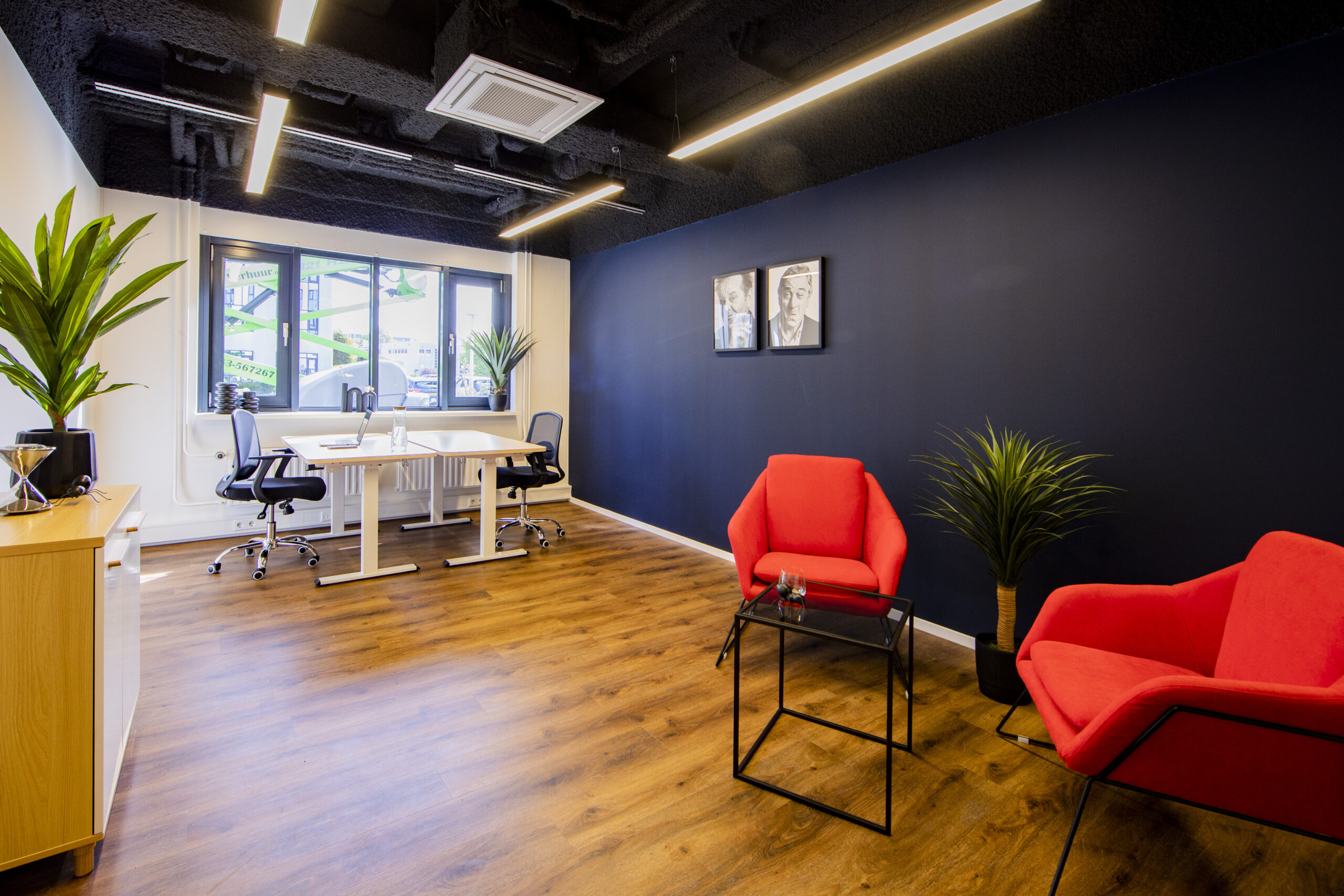 Modern kantoorinterieur met houten vloer, rode fauteuils, een met planten ingerichte werkruimte en kunst aan de muur.