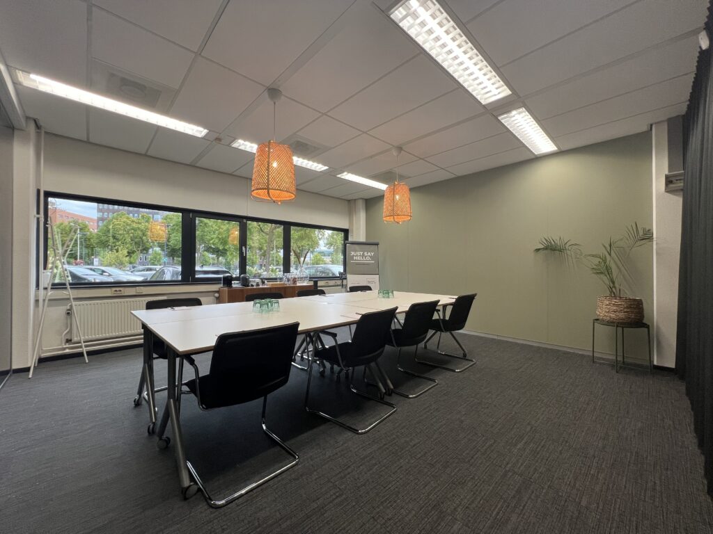 Vergaderruimte met een lange tafel, zwarte stoelen, grote ramen en hanglampen aan het plafond.