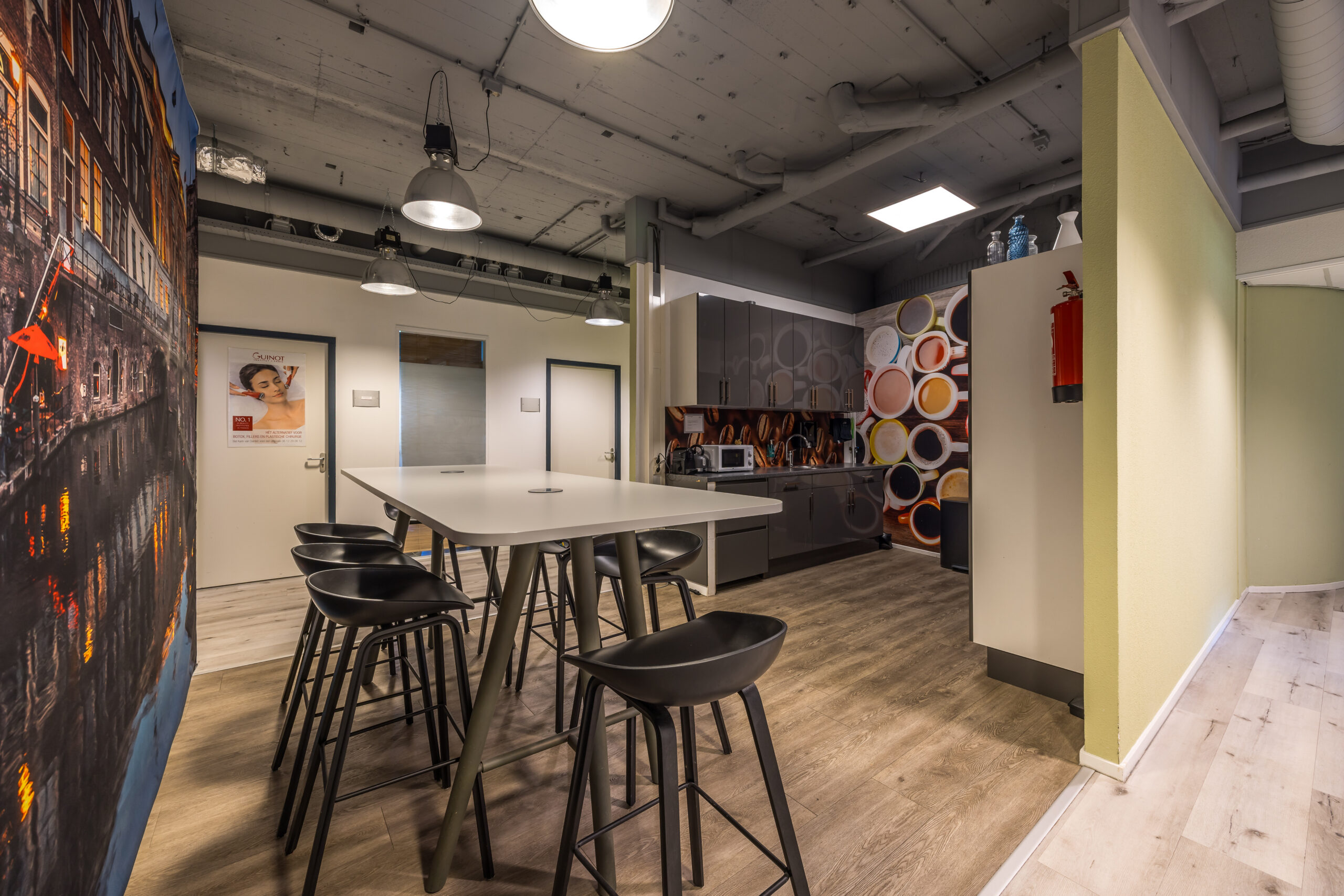 Keukenruimte in een modern kantoor met hoge tafels en barkrukken, kleurrijke wanddecoraties en een kitchenette met apparatuur.