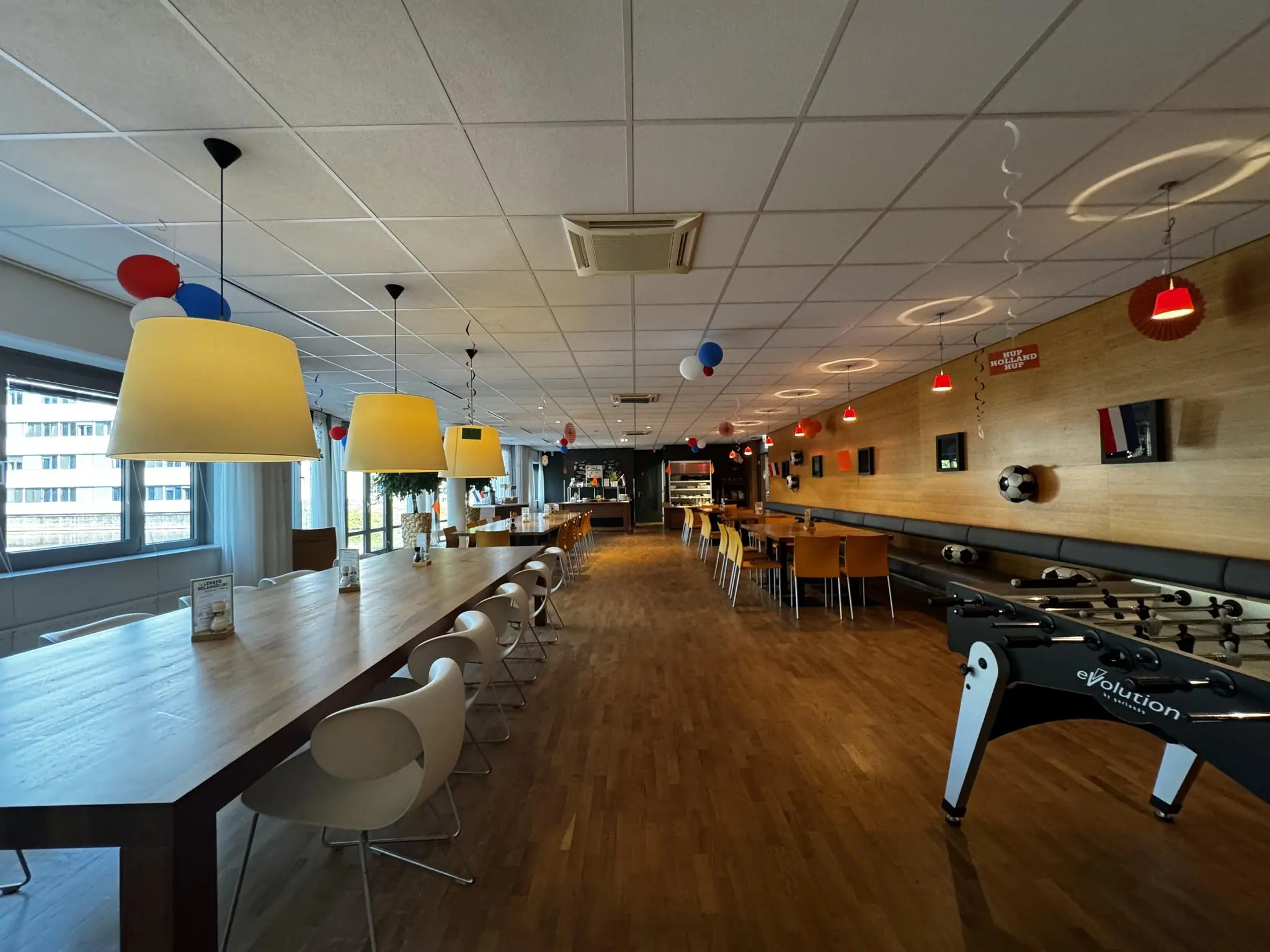 Moderne bedrijfskantine aan de Softwareweg met lange houten tafels, sfeerverlichting, een tafelvoetbaltafel en oranje feestdecoratie.
