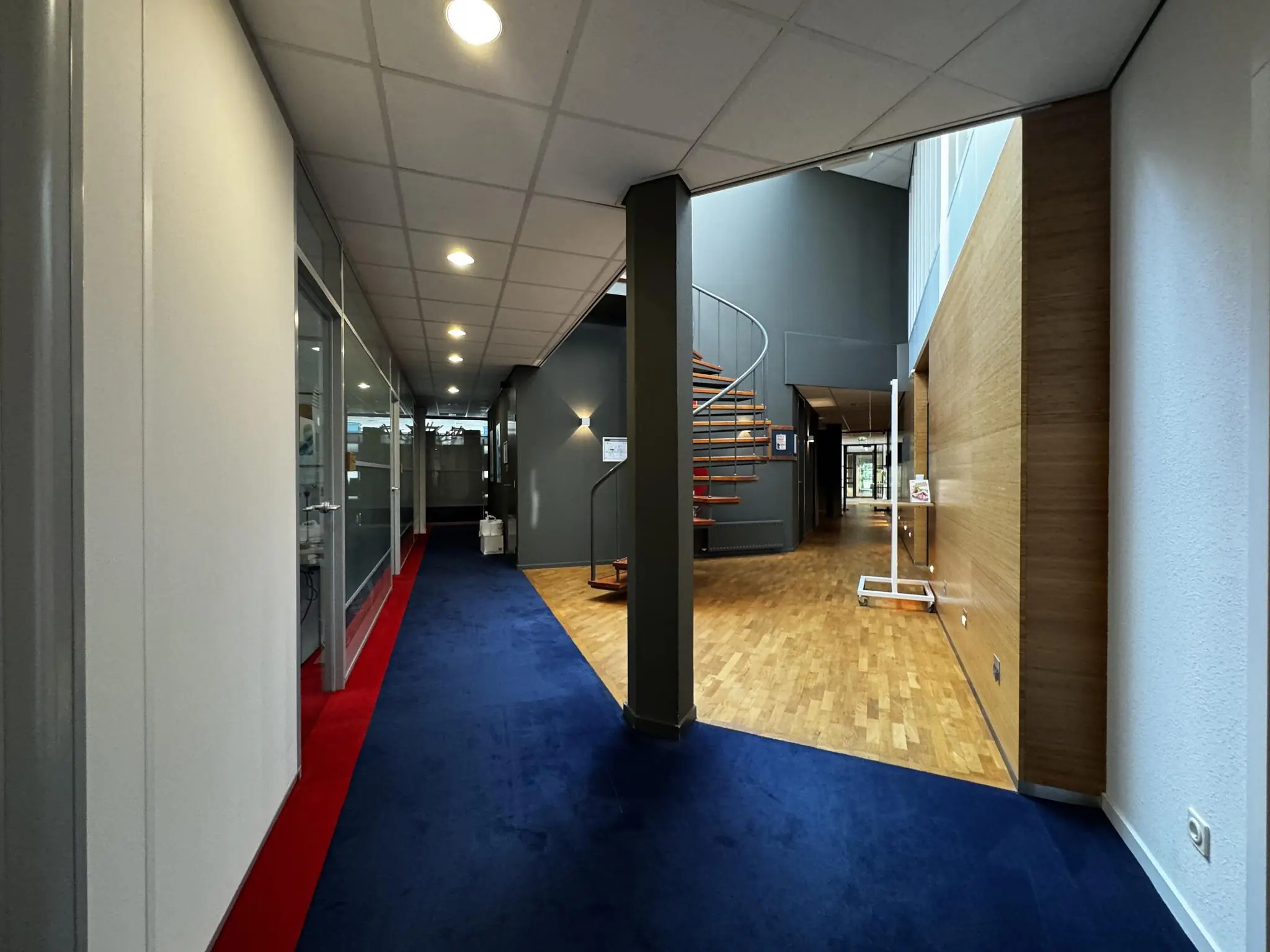 Moderne kantoorhal met blauwe en rode vloerbedekking, glazen wandkantoren en een houten wenteltrap.